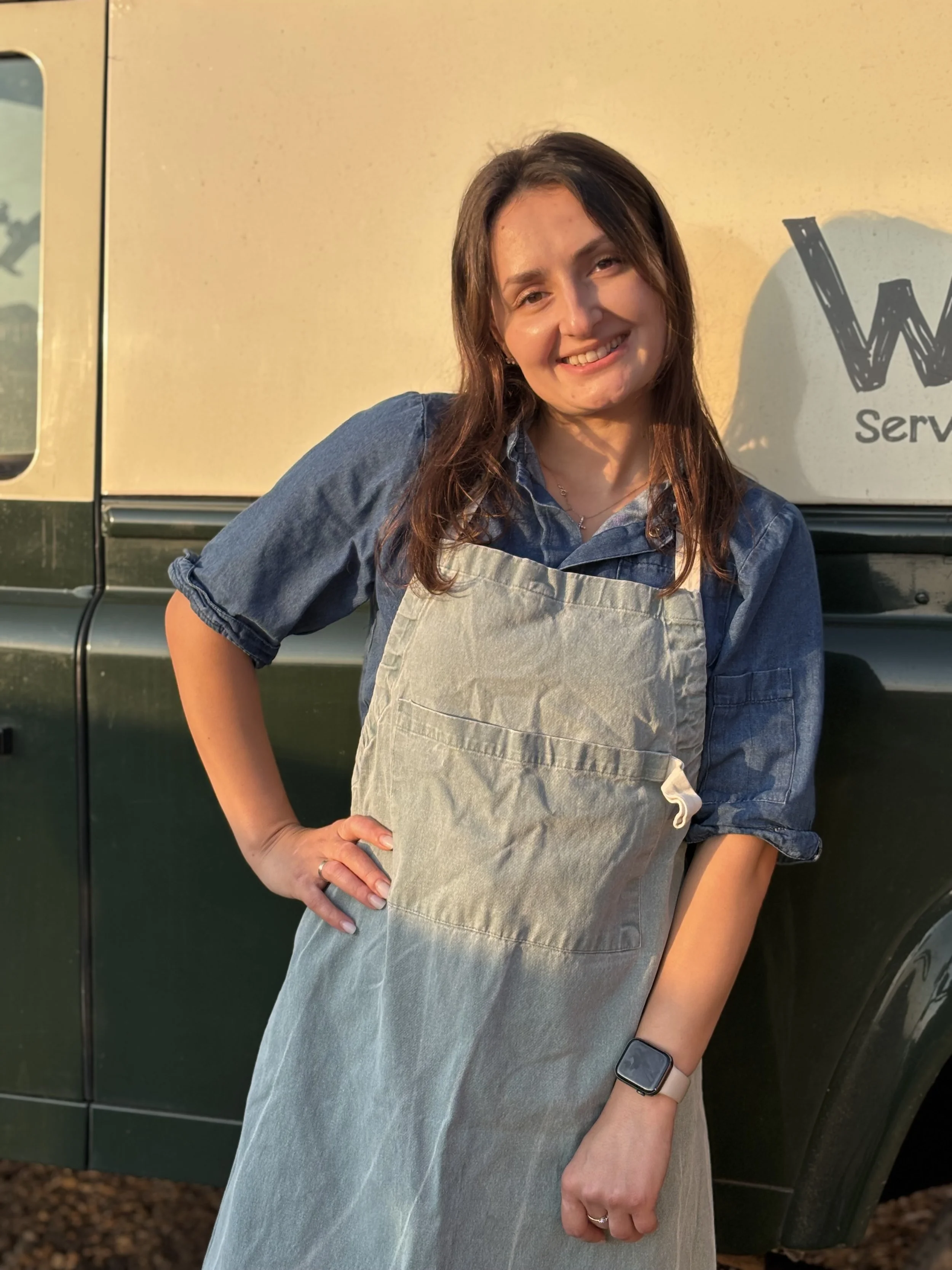 A woman with long brown hair wearing a blue denim shirt and a beige apron, smiling and standing in front of a yellow wall and a green vehicle with a partial sign that reads 'W... Service'.