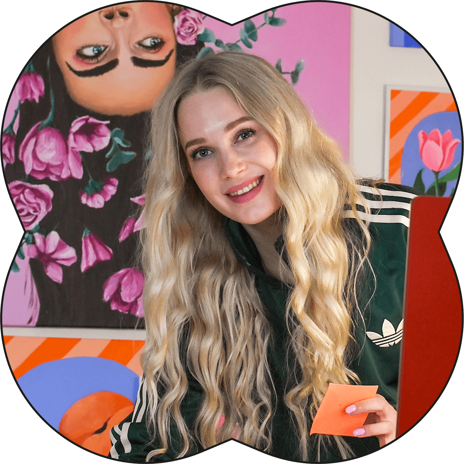 Nuori nainen hymyilee, pitkät laineiset hiukset, vihreä Adidas-uimapuku, istuu edessä, taustalla värikkäitä tauluja, yksi taulu sisältää pinkkejä kukkia ja toinen on abstrakti, pinkeillä ja oransseilla väreillä.