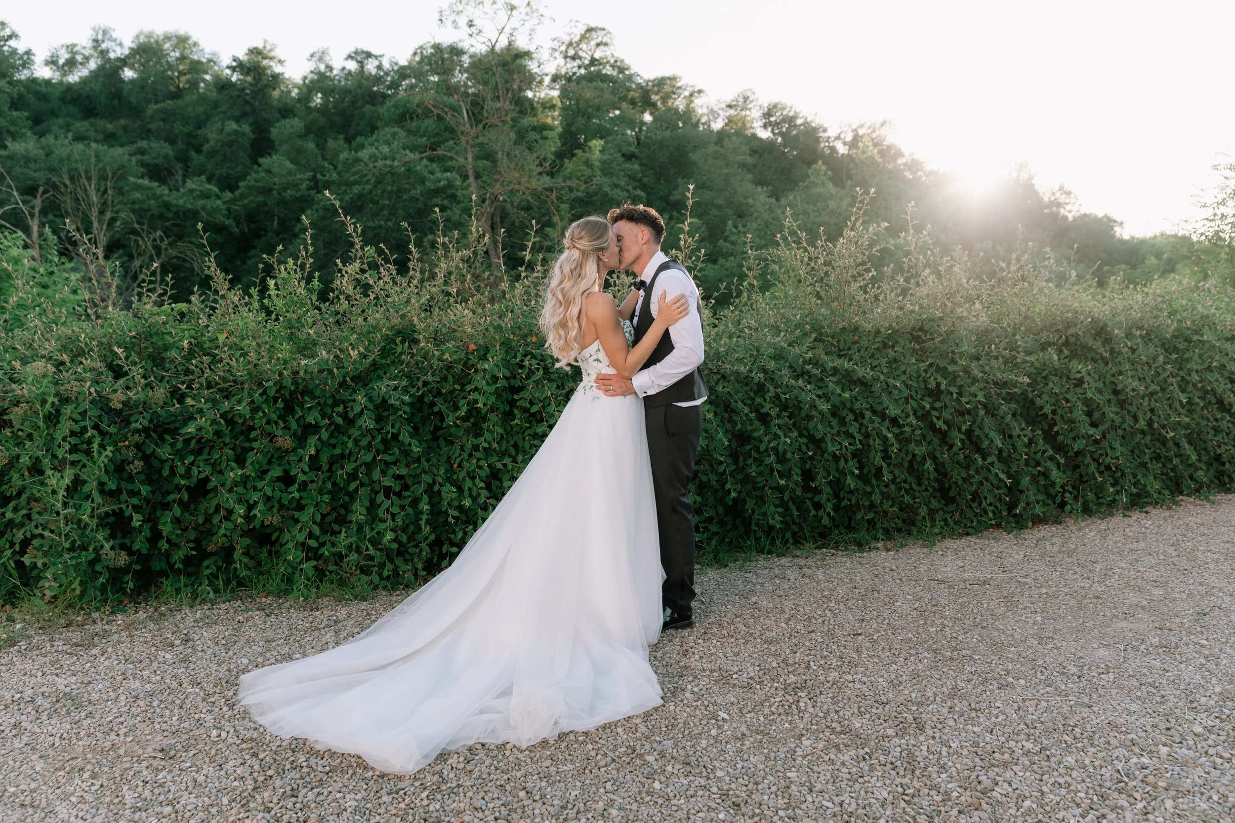 A bride and groom kissing outdoors during sunset, surrounded by greenery with a gravel path in the foreground.