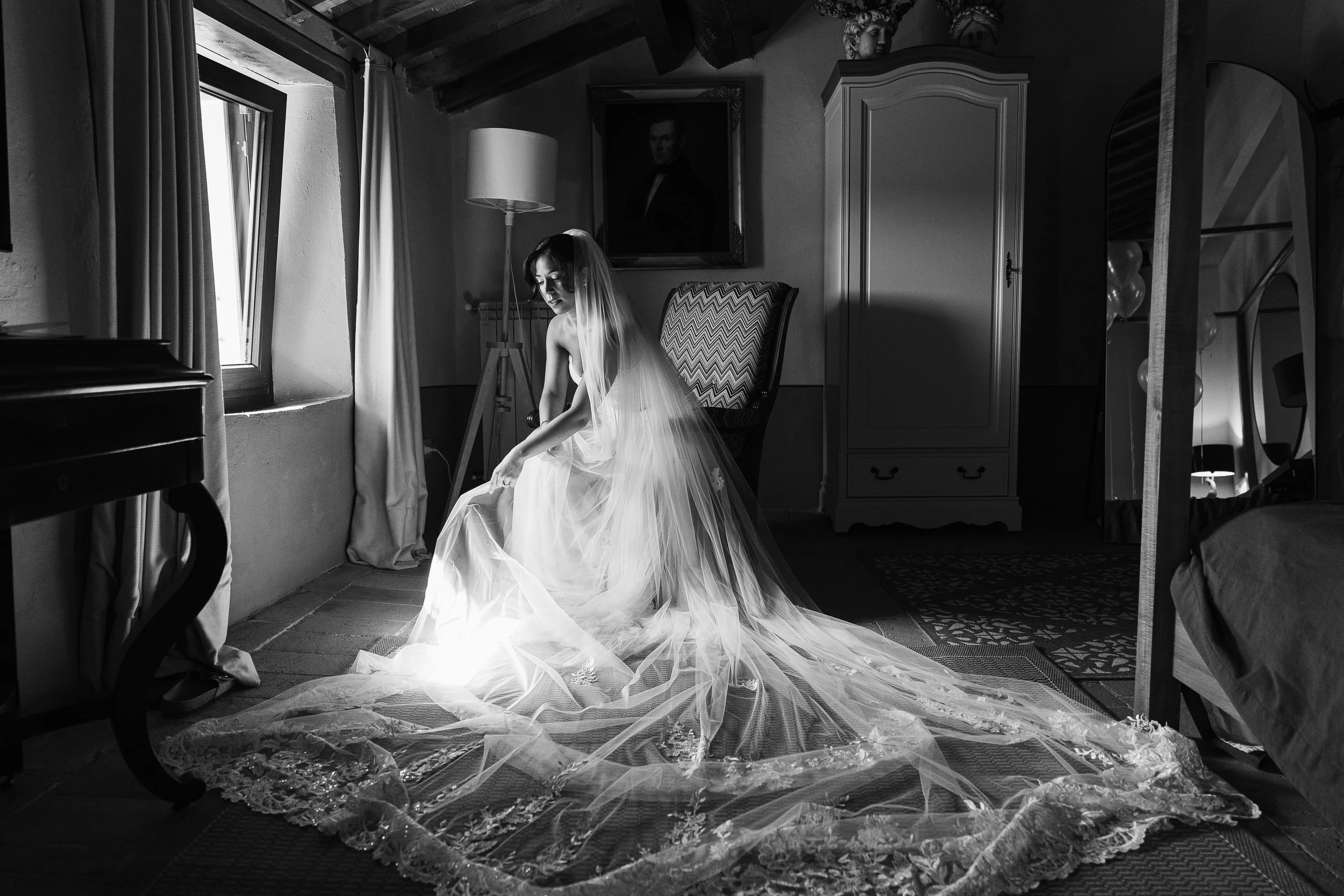 A bride sitting in a room with rustic wooden ceiling beams, she is wearing a wedding dress and veil, with a long train spread out on the floor, and natural light coming from a window on the left.