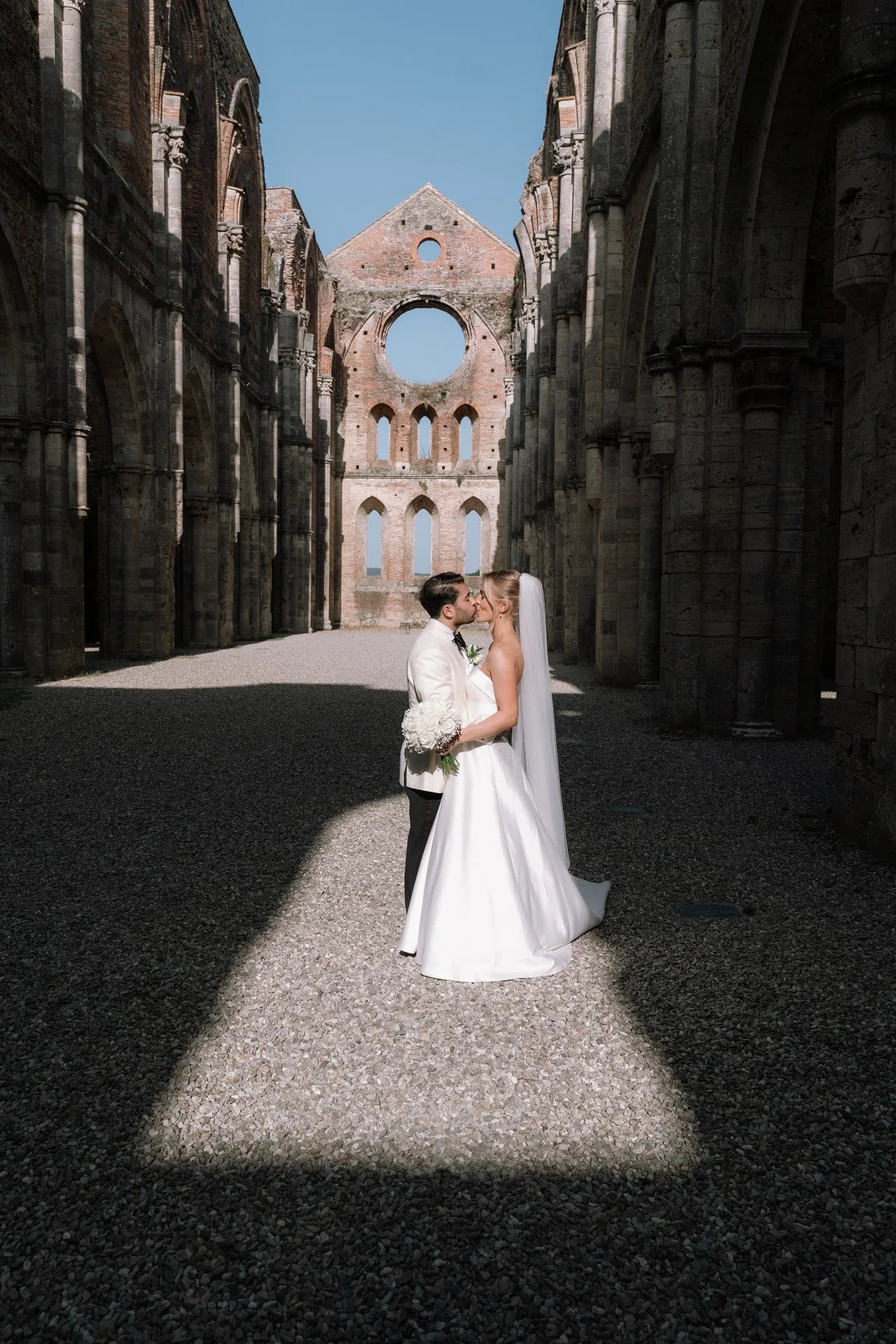 A bride and groom are standing face to face in front of the ruins of an old stone church. The groom is in a white tuxedo, and the bride is in a traditional white wedding gown with a veil. They are about to kiss, with a clear blue sky visible through 