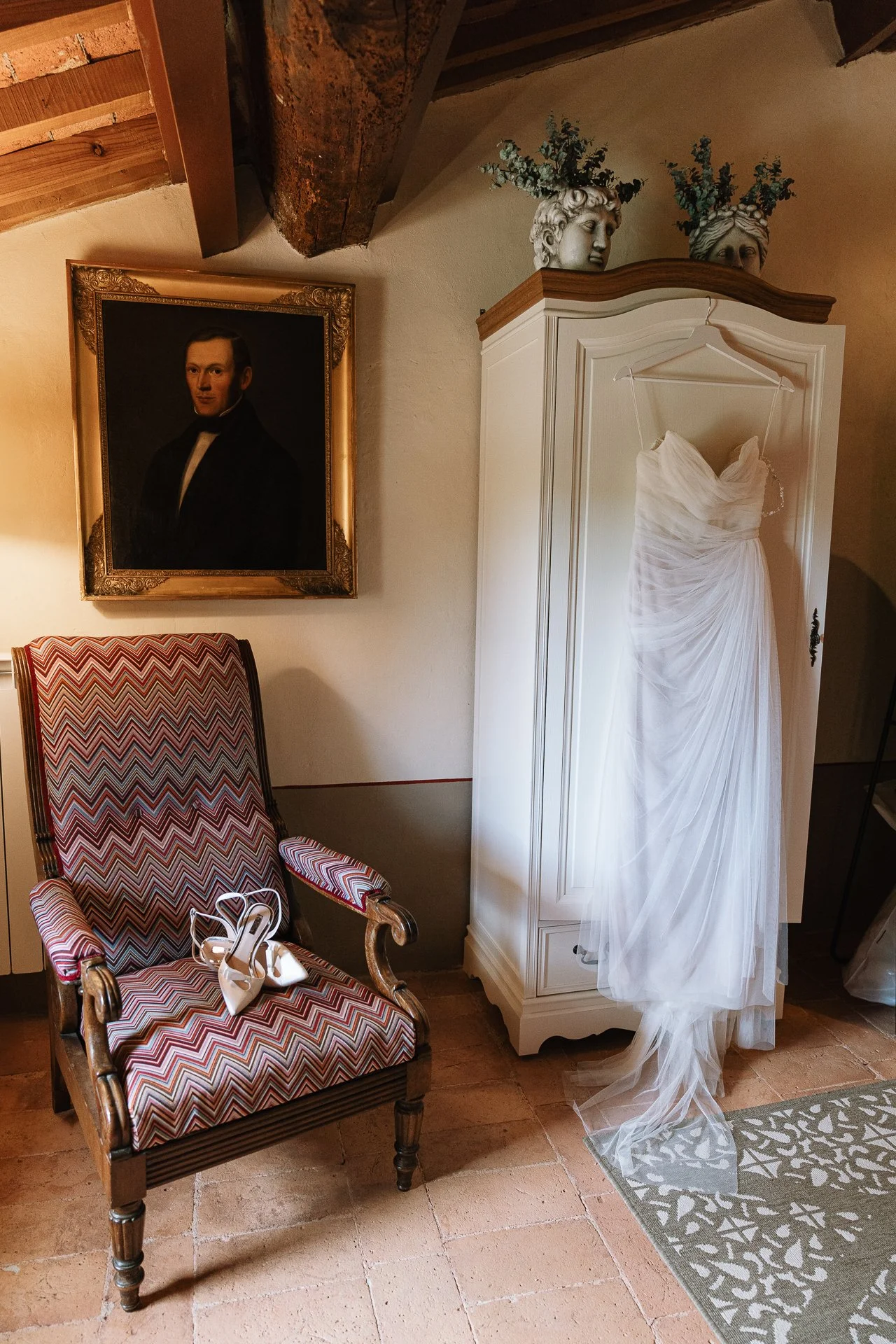 A wooden armchair with a colorful zigzag-patterned upholstery, a pair of white high-heeled shoes with an ankle strap resting on the seat. To the right, a white wardrobe with a wedding dress hanging on a hanger. On top of the wardrobe, two decorative 