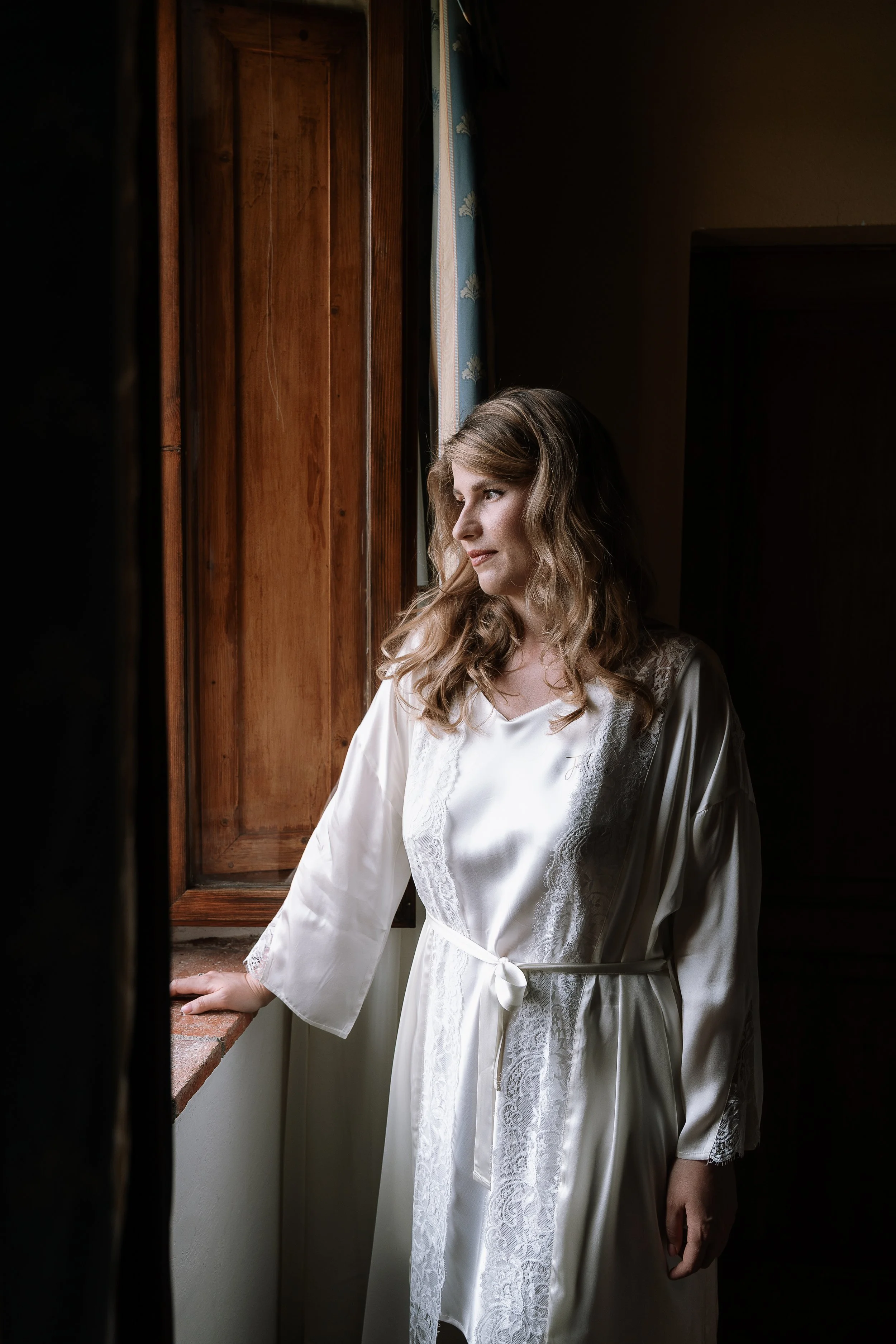 A woman with wavy blonde hair wearing a white satin robe with lace details, standing by a window with wooden shutters, looking outside with a calm expression.
