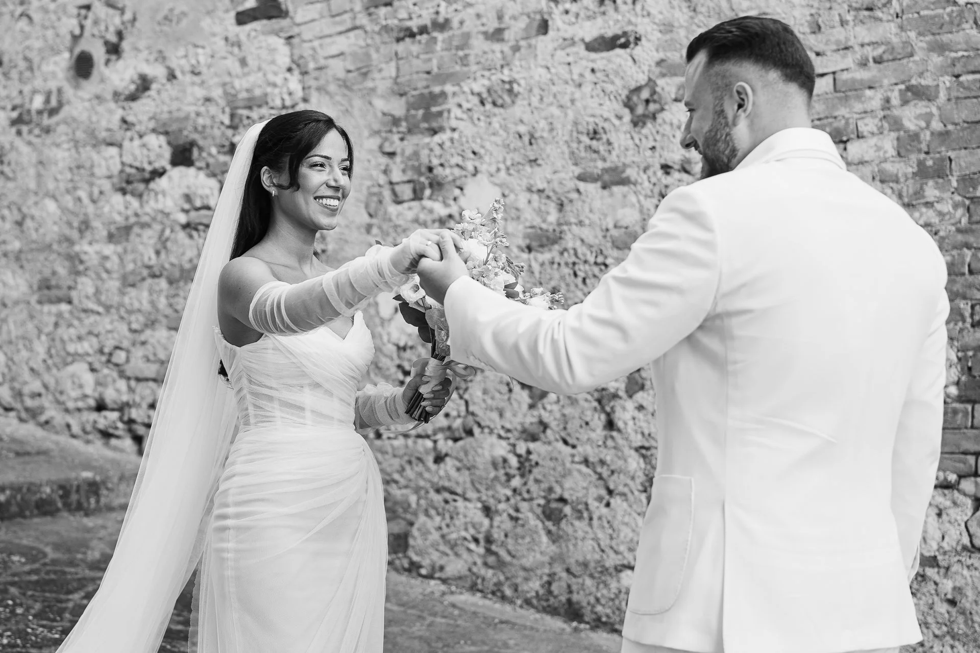 A bride in a wedding dress holding a bouquet of flowers and smiling at a groom, who is dressed in a white suit, during their wedding ceremony outdoors against a brick wall.