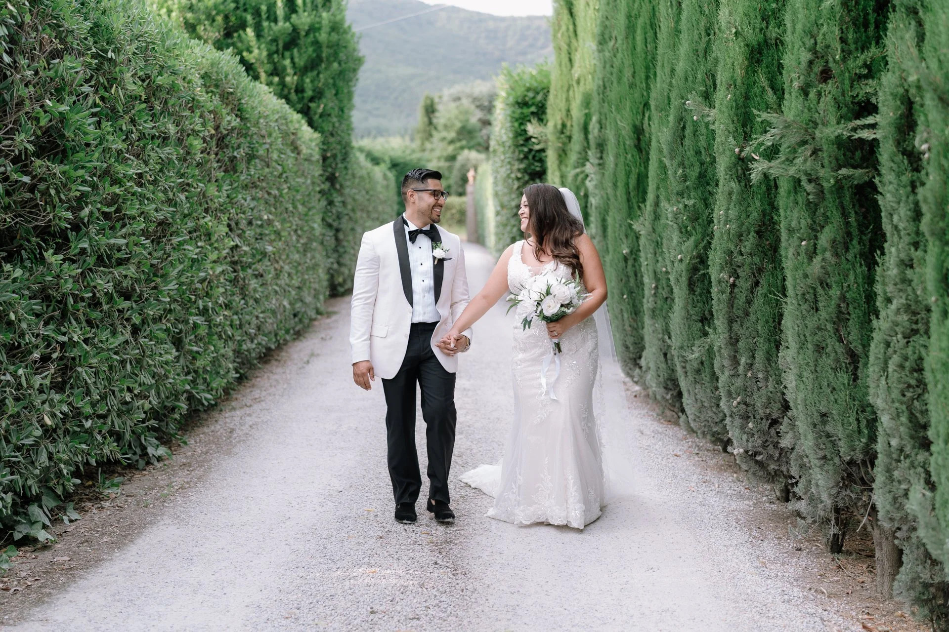 A bride and groom standing in front of a wooden door, looking at each other and holding a bouquet of white flowers.