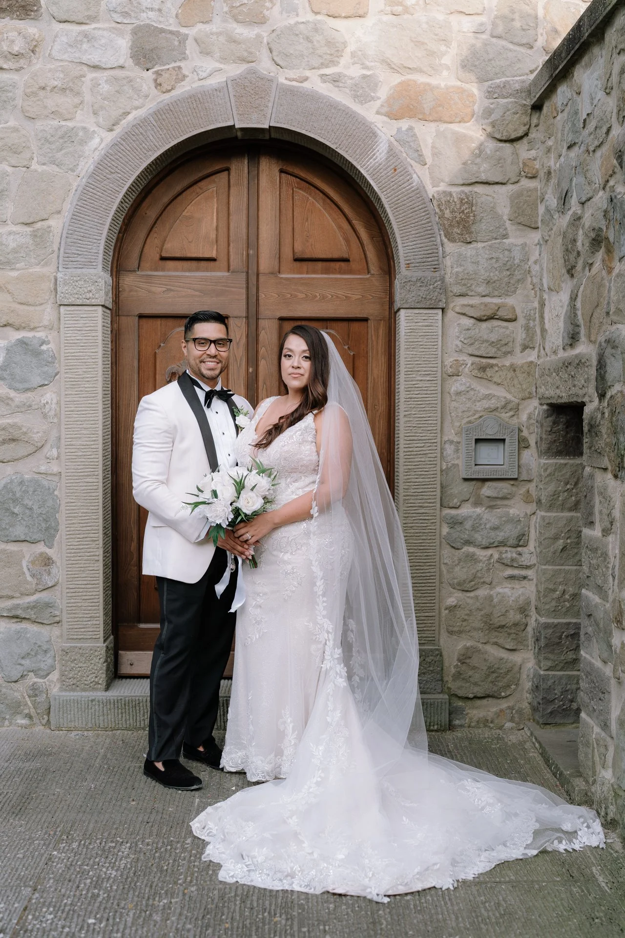 A bride and groom standing together in front of a stone wall and wooden door on their wedding day. The groom is wearing a white tuxedo jacket with black lapels, black pants, and a black bow tie, holding a bouquet of white flowers. The bride is in a w
