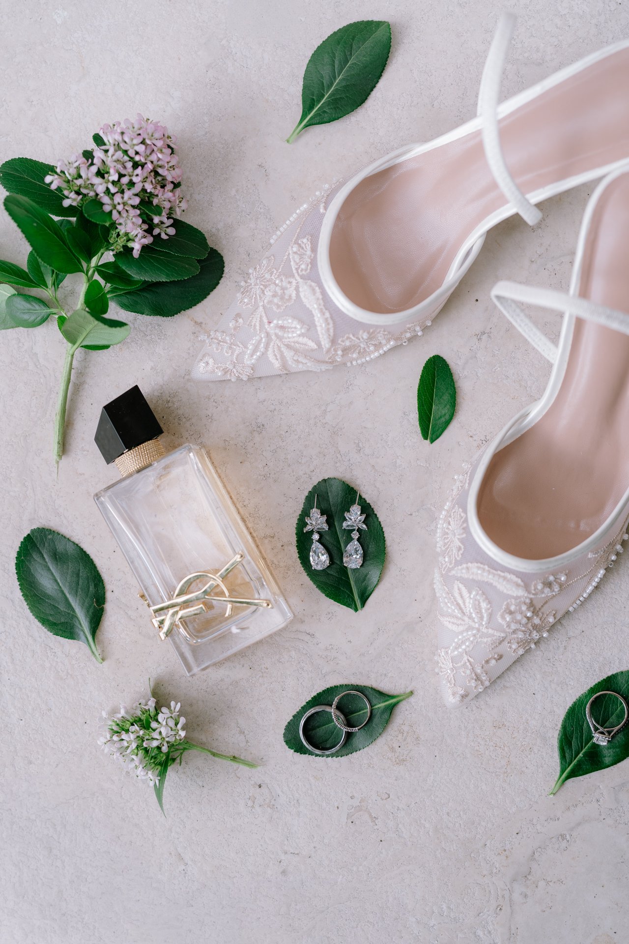 Pink wedding shoes with lace detail, surrounded by green leaves, flowers, jewelry including rings and earrings, a perfume bottle, and a small display case on a light textured surface.