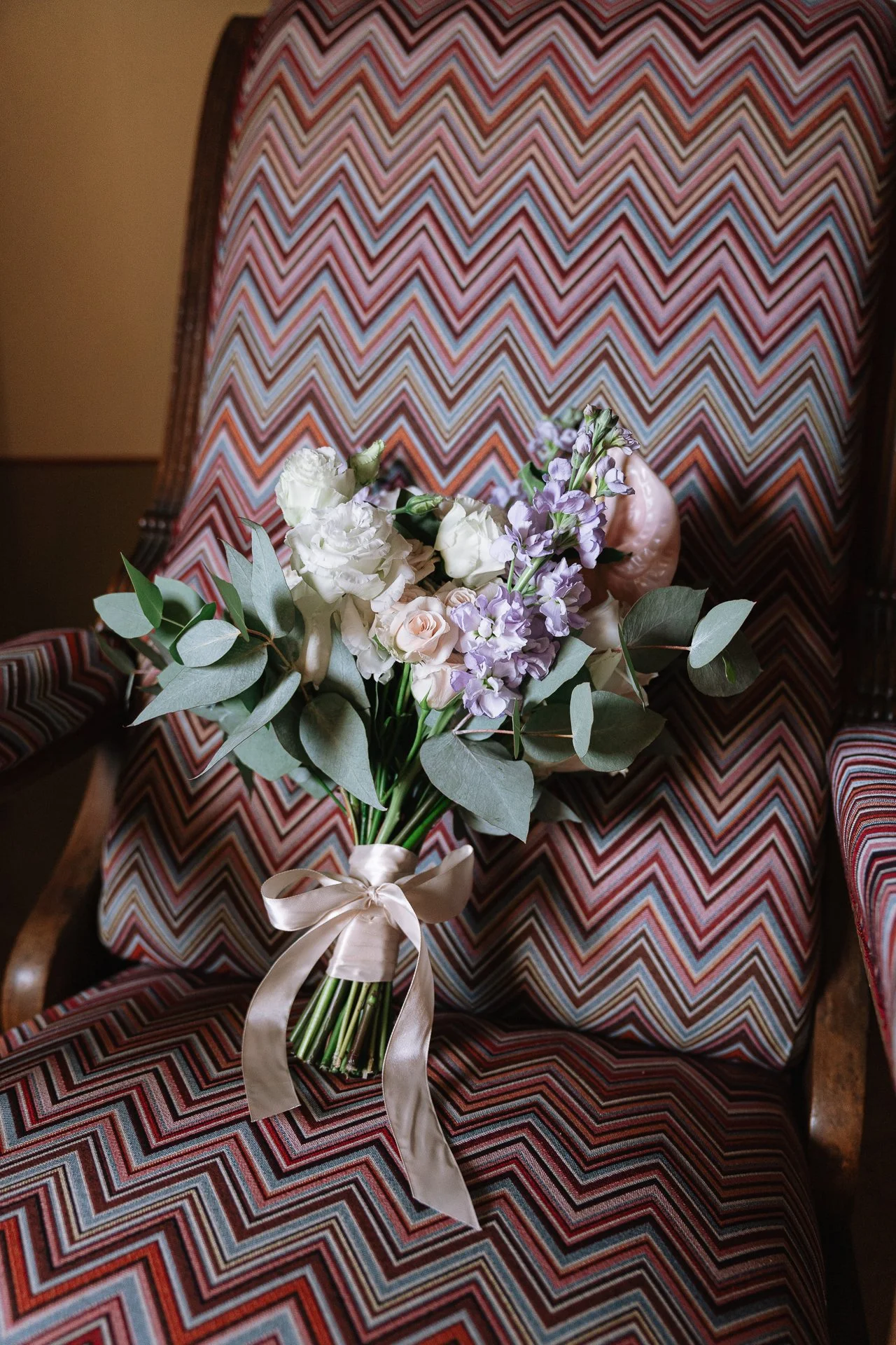 A bouquet of white and purple flowers with green leaves tied with a silver ribbon, resting on a pink, red, blue, and white zigzag patterned chair.