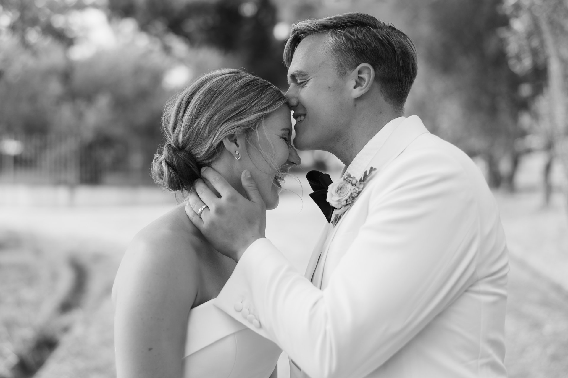 Black and white photo of a couple in wedding attire, smiling and embracing outdoors, close-up of their faces touching, joyful moment.