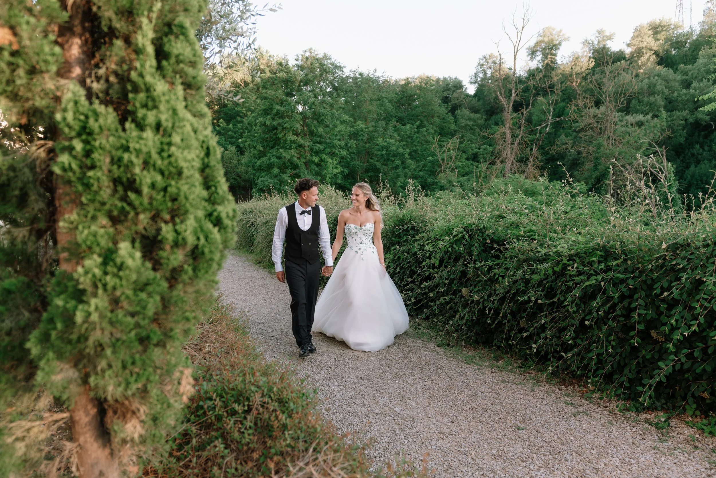 A bride and groom walking hand in hand along a garden path, surrounded by lush greenery and trees at Agriturismo Antiche Dimore di Poggianto.