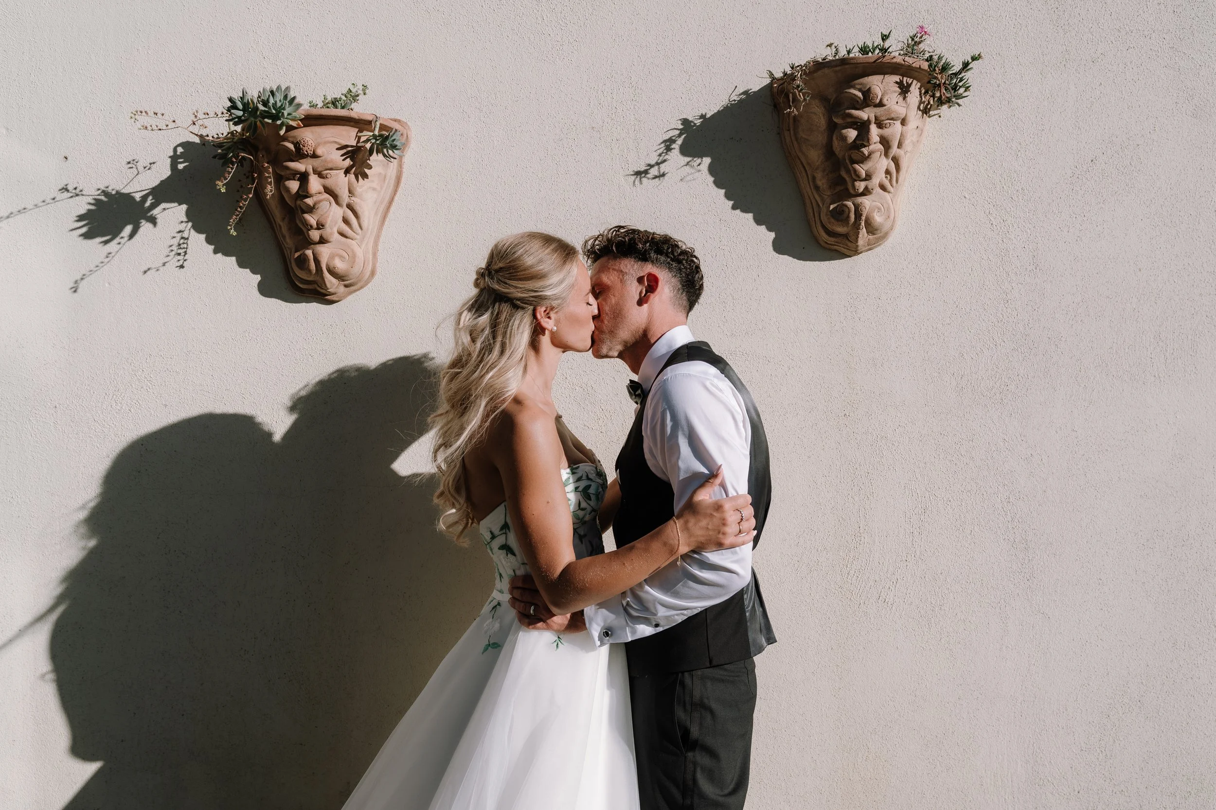 A couple dressed in formal wedding attire sharing a kiss against a white wall with decorative wall planters featuring greenery and sculptures that cast shadows.