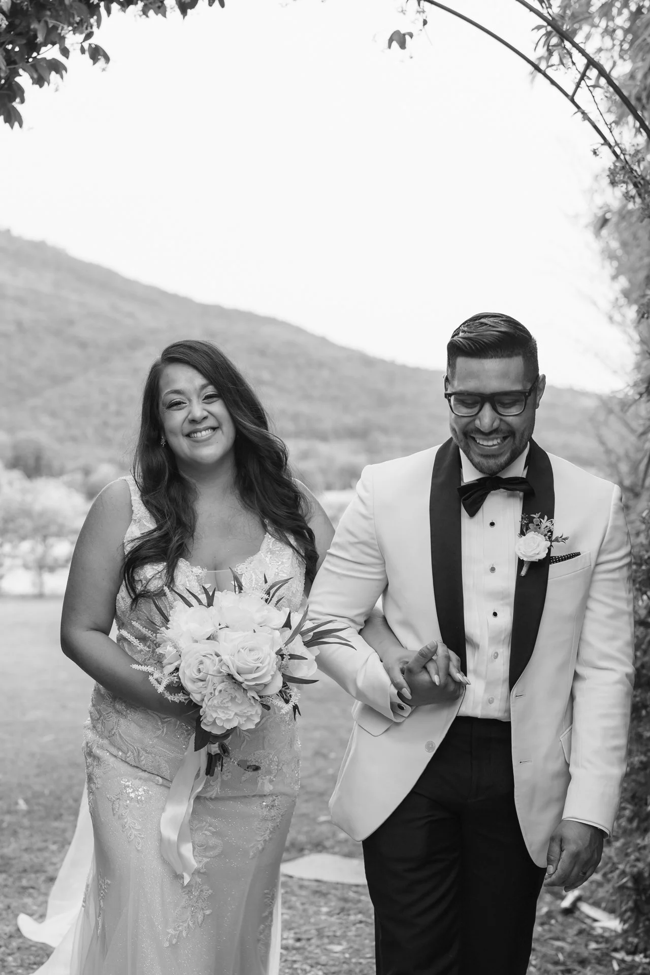A black and white photo of a smiling bride and groom walking outdoors hand in hand, with trees and hills in the background at La Corte dei Papi, Cortona, Tuscany.