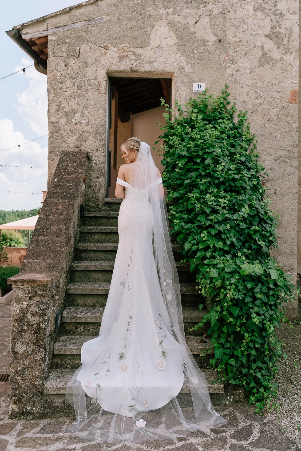 A bride in a white wedding gown with floral embroidery stands on outdoor stone stairs, facing away, next to a large green ivy plant and an old stone building with a sign that reads '9 PANORAMICA'.