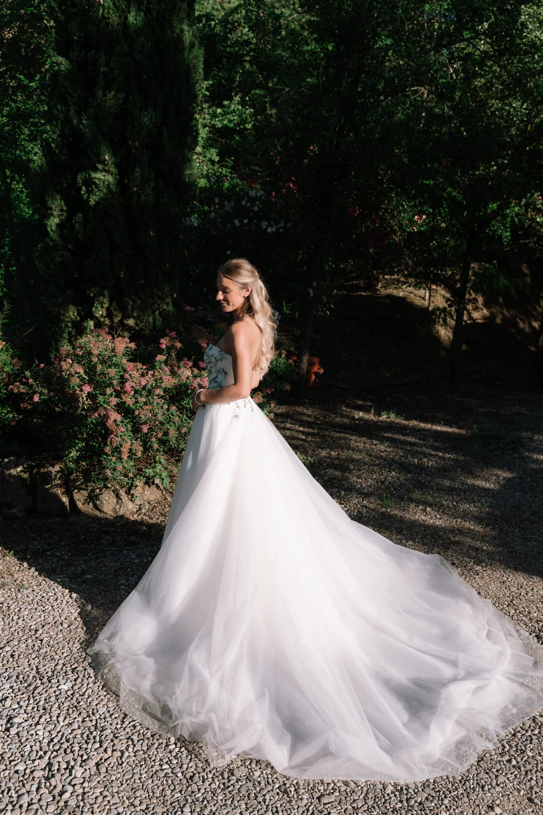 A woman in a white strapless wedding dress standing outdoors on a gravel path surrounded by green trees and bushes, with sunlight highlighting her and her dress.