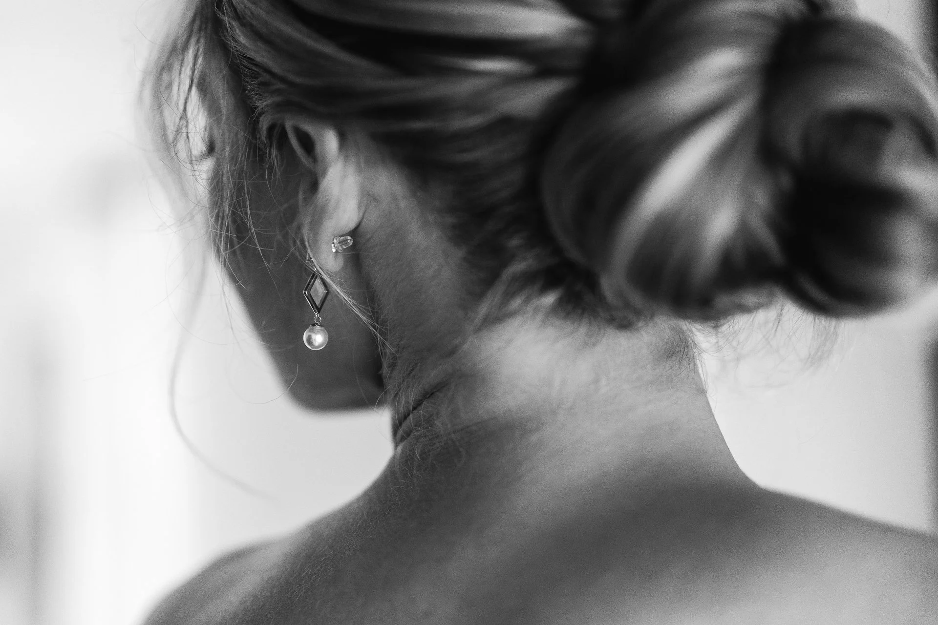 Close-up black and white photo of a woman's ear with multiple earrings, including a pearl and a geometric-shaped earring, her hairstyle is swept to the side.