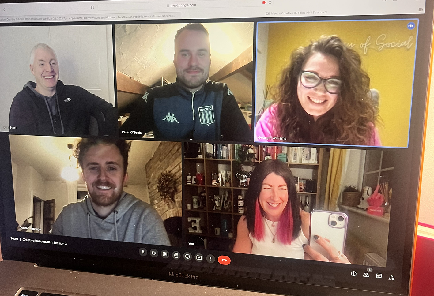 A screenshot of five people participating in a video call on a laptop, smiling and looking at the camera. The participants are in various indoor settings.