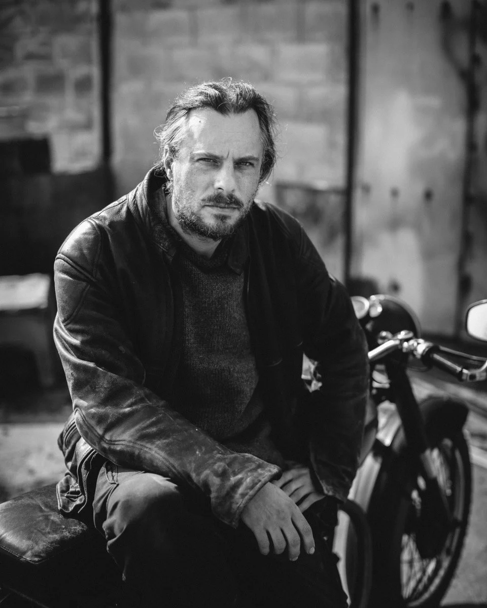 A man with a serious expression, long hair, and a beard, wearing a leather jacket, sitting on a bench in a dimly lit room with a motorcycle beside him, in black and white.