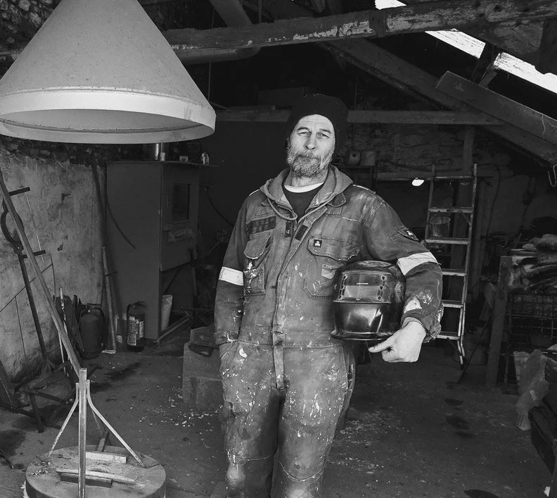 A man standing in a cluttered workshop holding a welding helmet, wearing a work jumpsuit with paint splatters, and a beanie, surrounded by tools and equipment.