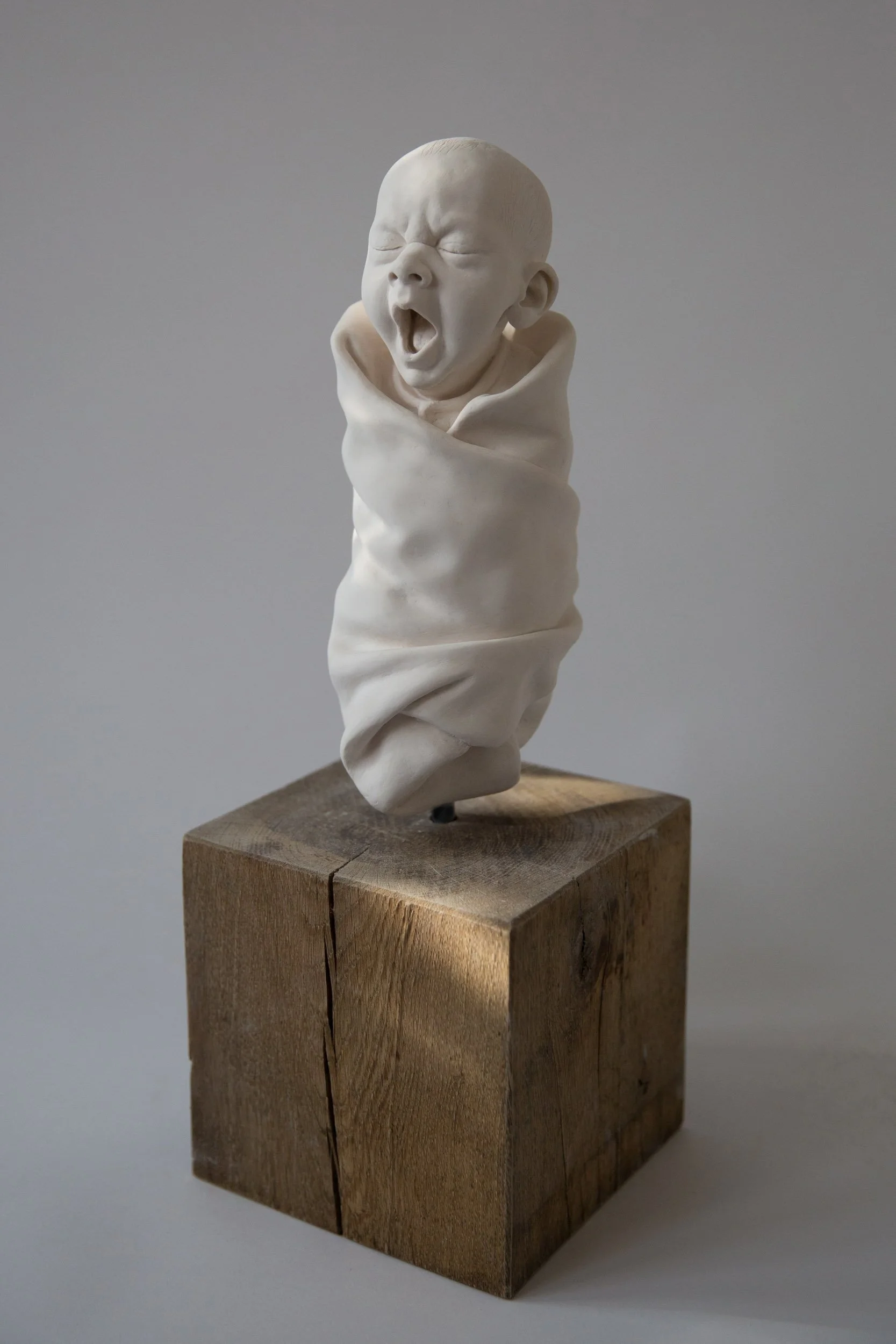 A sculpture of a yawning baby wrapped in a cloth, mounted on a wooden block.