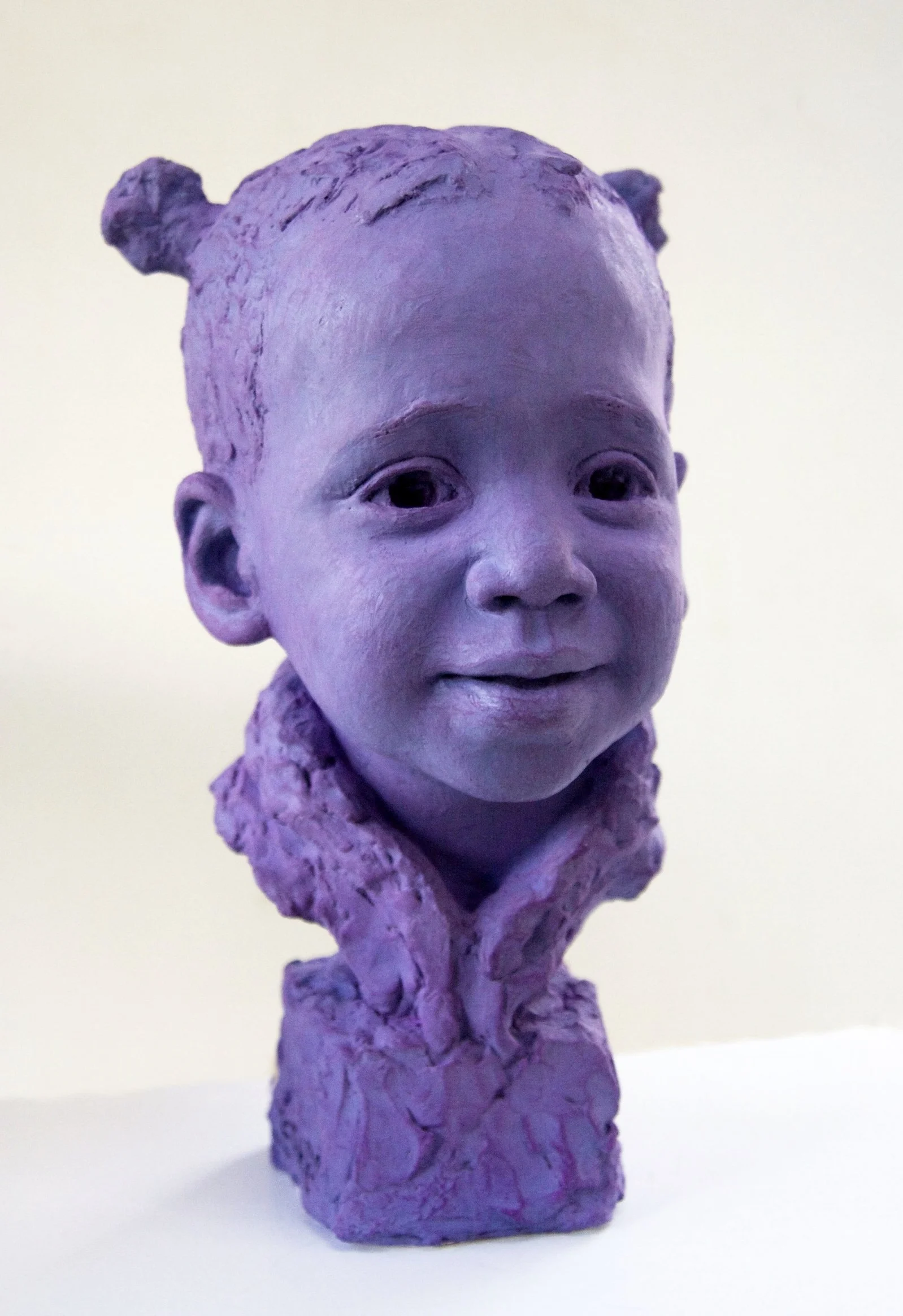 Sculpture of a young girl's face and shoulders painted purple, with textured hair styled in two buns, mounted on a purple textured base.