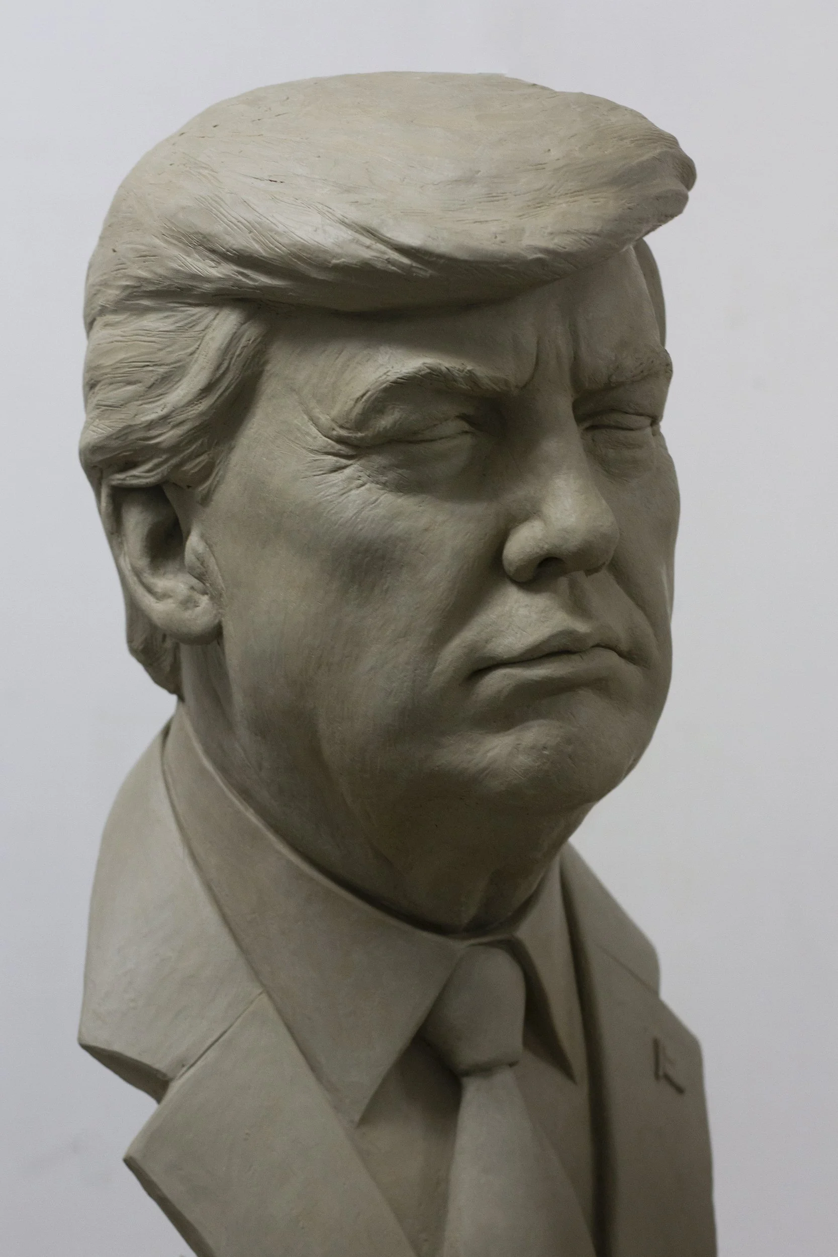 Close-up of a beige bust sculpture of a man with closed eyes, wearing a collared shirt, against a plain gray background.