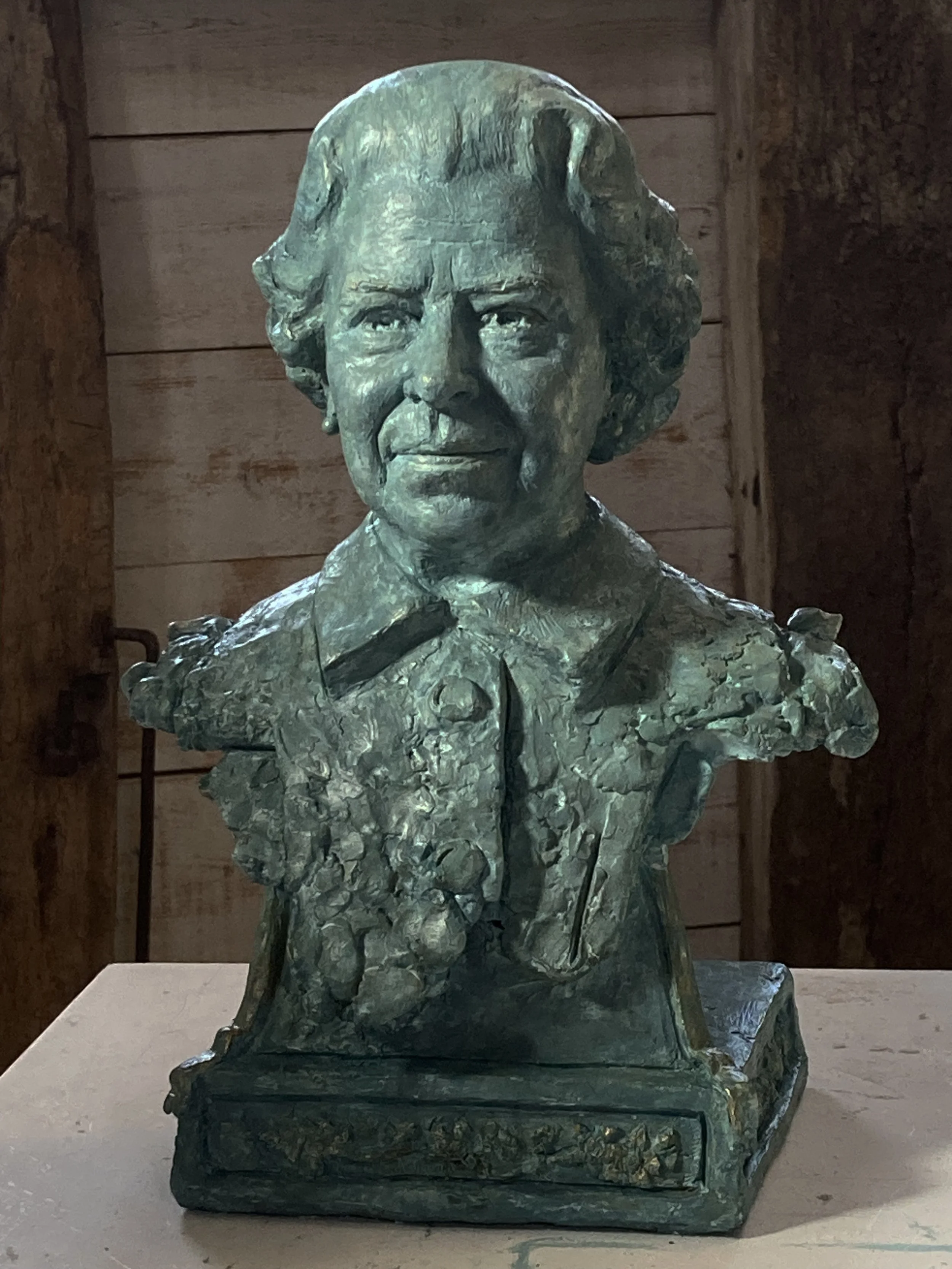 Bronze bust of a woman with curly hair, wearing a scarf or collar, displayed on a pedestal against a wooden wall background.