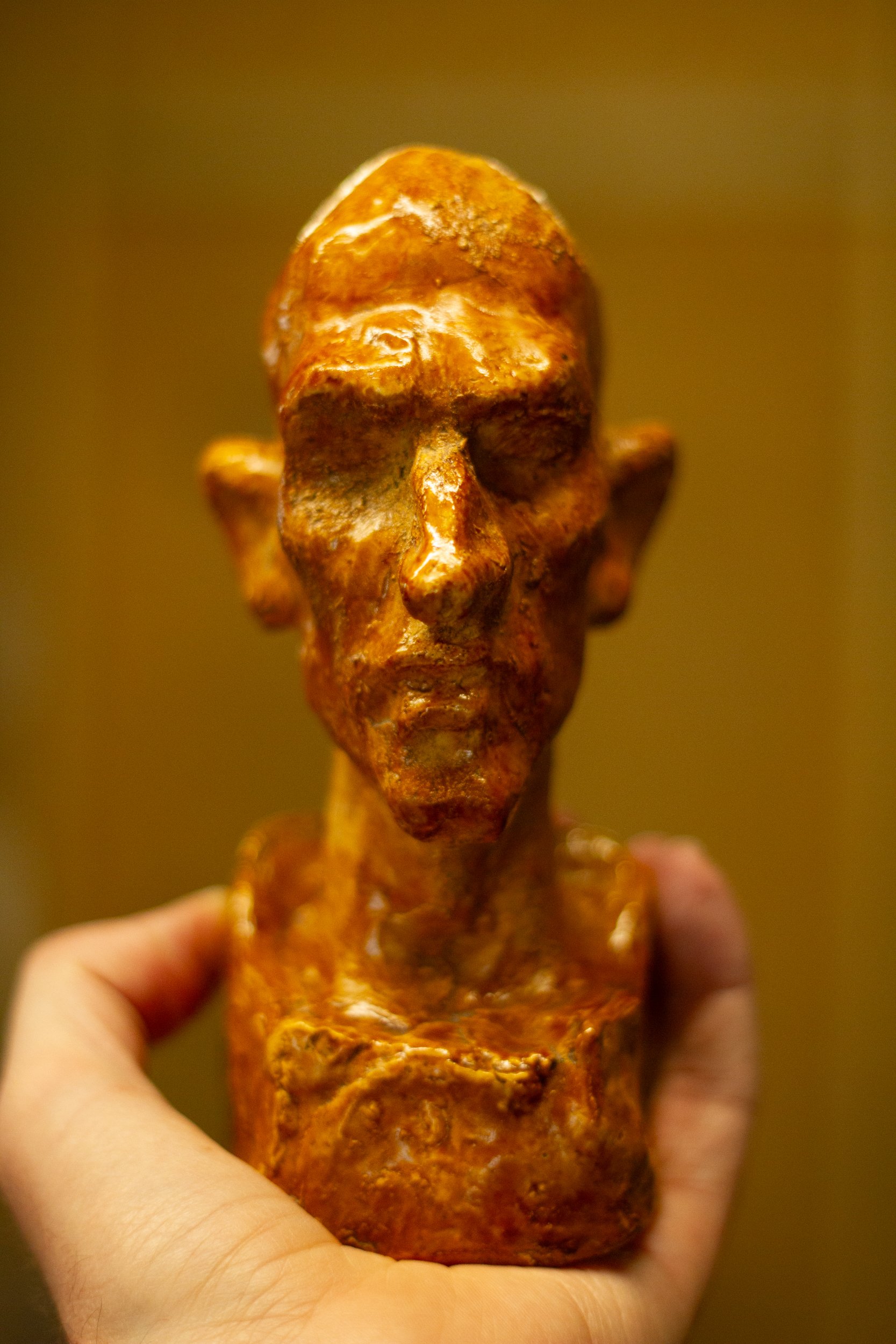 Close-up of a small sculpture of a human face held in a person's hand, made of metal with a bronze appearance.