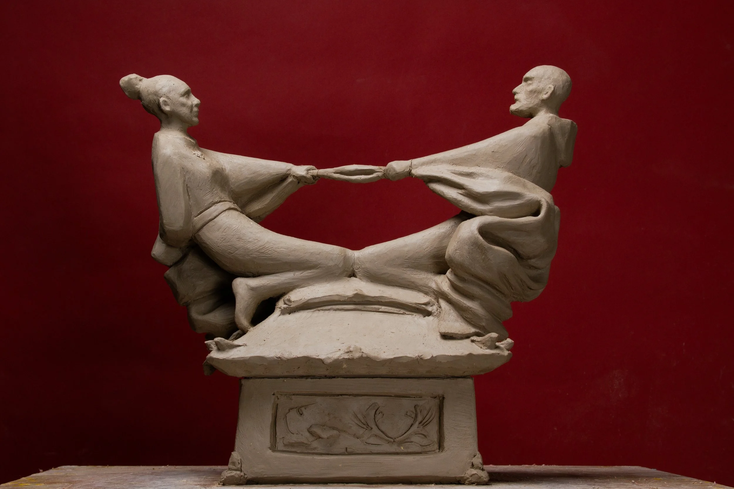 Sculpture of two people holding hands and sitting cross-legged on a base, with a red background.