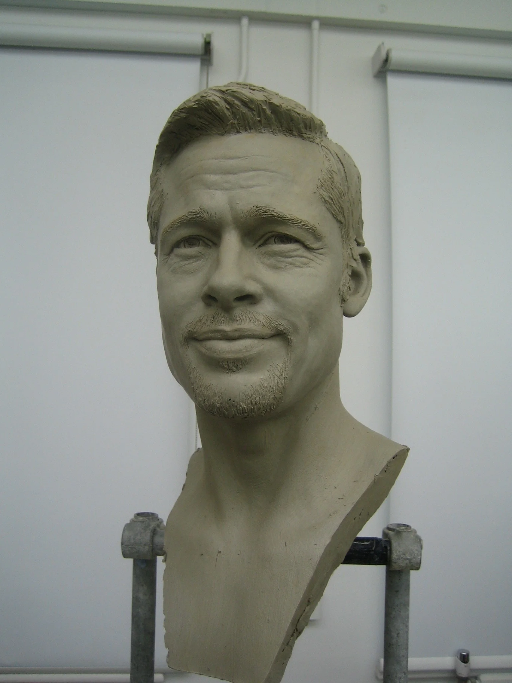 A clay sculpture bust of a man with short hair, a slight beard, and a content expression. The sculpture is set on a worktable in a studio.