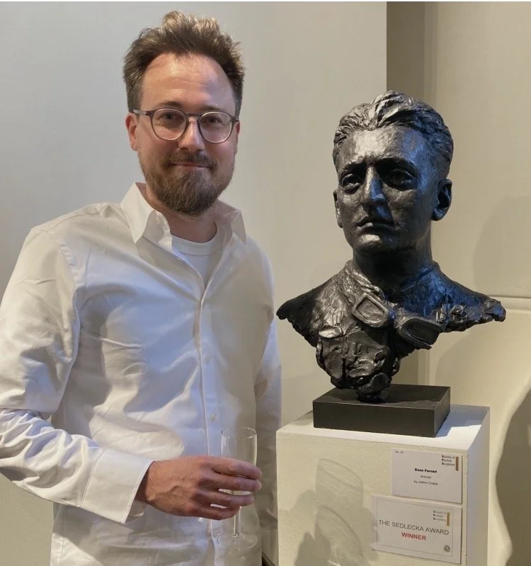 A man with glasses, a beard, and wearing a white shirt is standing next to a bronze bust sculpture of a woman with her hair slicked back, dressed in a military-style jacket with a bow tie. The man is holding a glass in his right hand, and there is a 