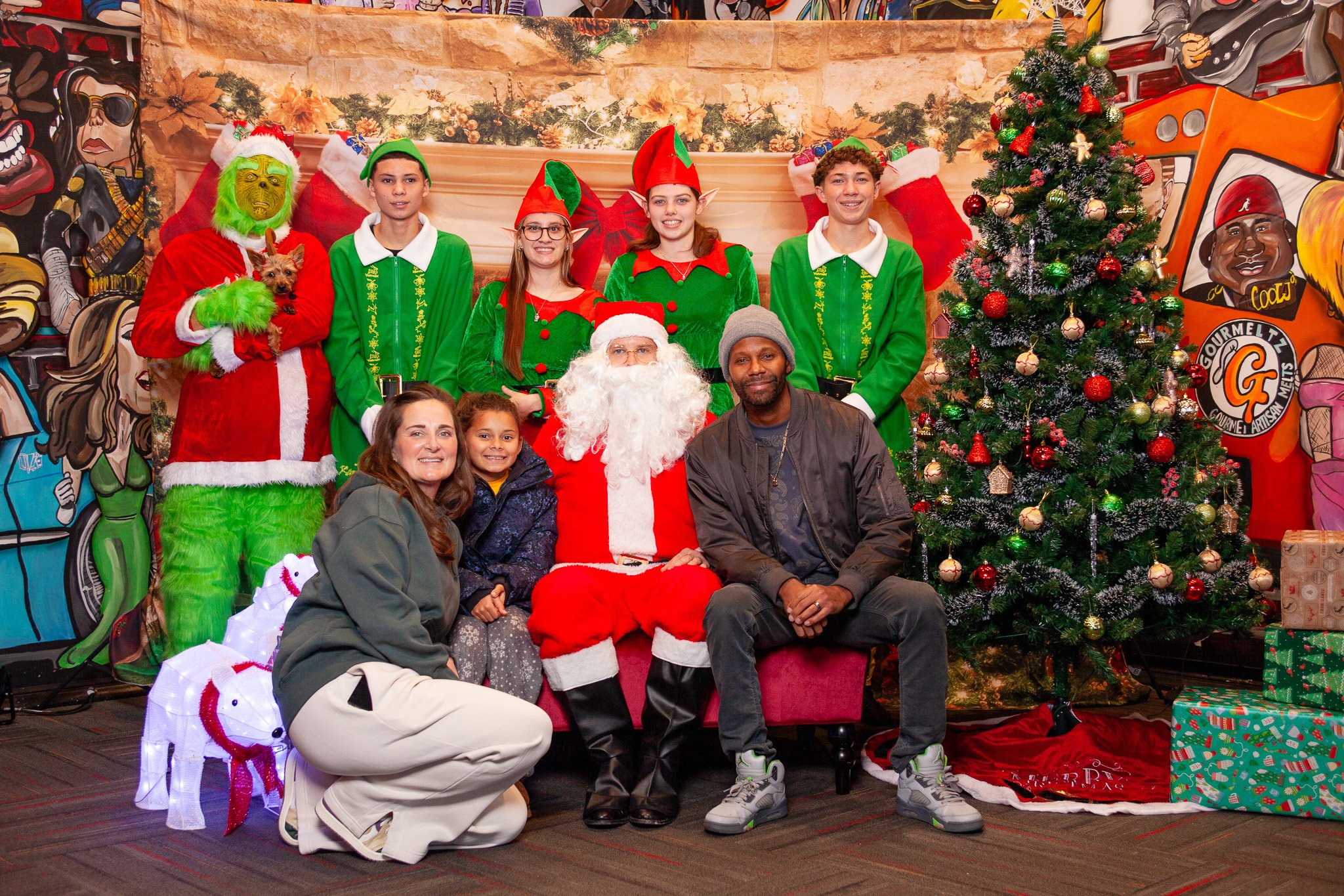 Group photo with Santa, elves, and people in festive costumes by a Christmas tree.