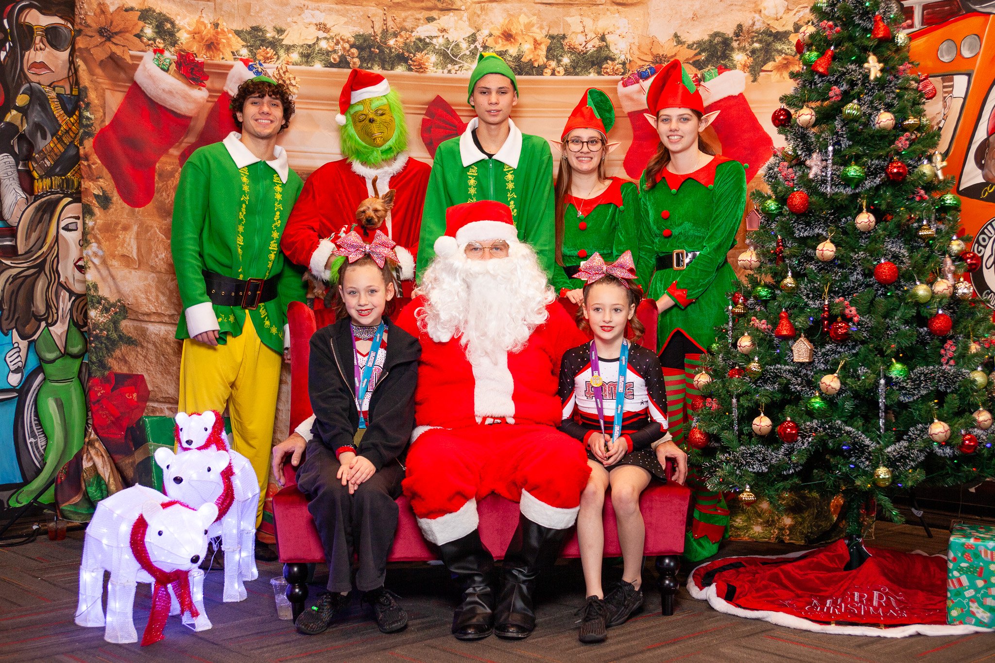 Group photo with people dressed as Santa Claus, elves, and a character resembling the Grinch, along with two children. They are posing beside a Christmas tree. Decorations include stockings, presents, and lighted polar bear figures.
