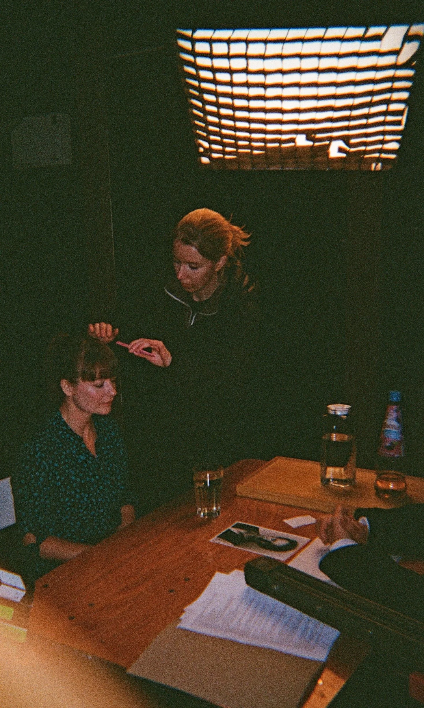 A woman getting her hair styled at a salon while sitting at a wooden table with papers, a pen, a water glass, and a water bottle. Another person’s hand is visible, likely a client or another stylist.