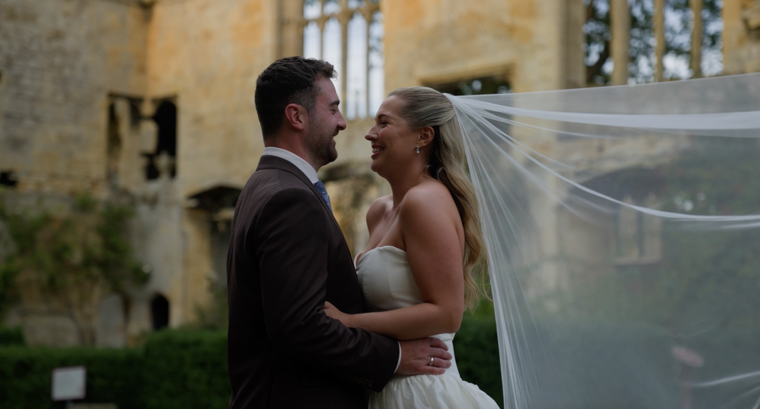 A couple, dressed in wedding attire, smiling and facing each other outdoors during their wedding, in front of an old stone building with greenery.