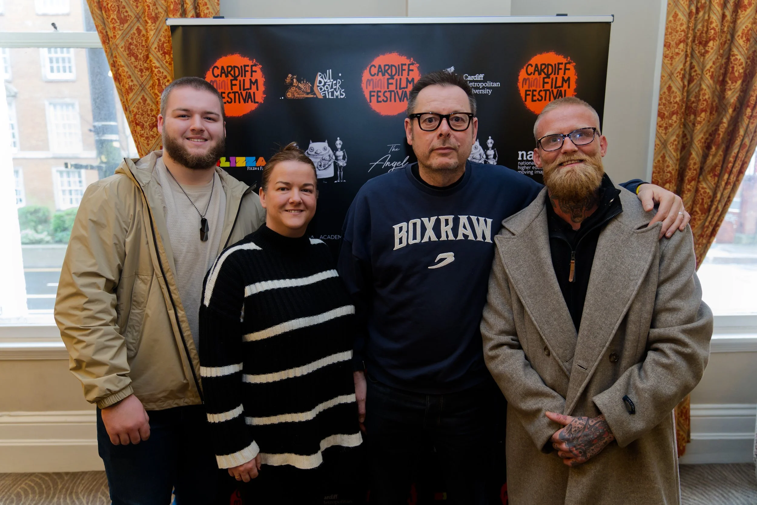 Group of five people standing in front of a black backdrop with logos, indoors with windows and curtains visible.