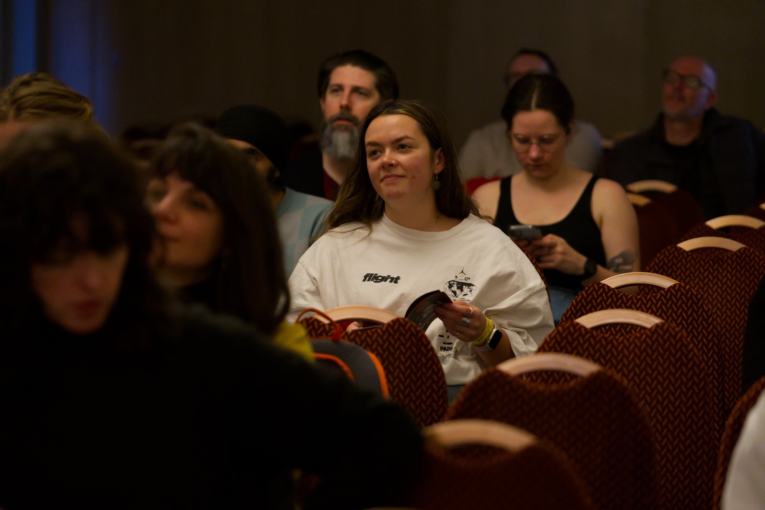 Audience members seated in rows at an indoor event, some looking at their phones, woman in the center wearing a white shirt with text and graphics, holding a booklet.
