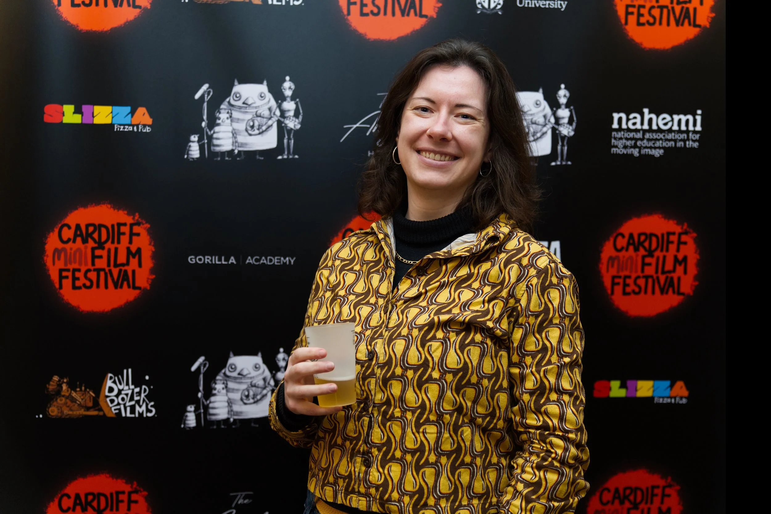A smiling woman holding a clear cup with a yellow beverage, standing in front of a black backdrop with logos for Cardiff Mini Film Festival and other sponsors.