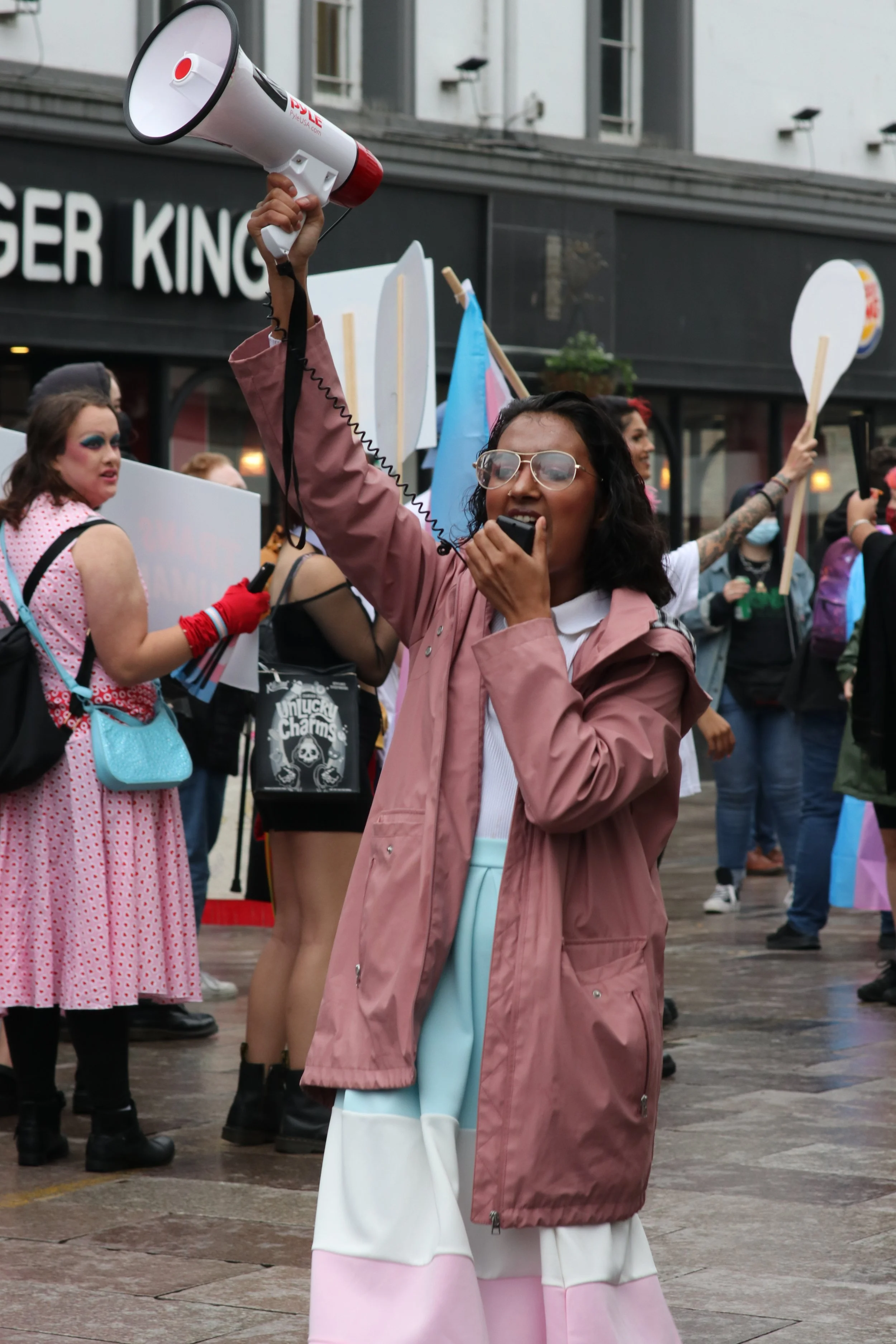 Woman speaking into a megaphone at a crowded protest or rally, surrounded by other protesters holding signs and flags, in front of a Burger King storefront.