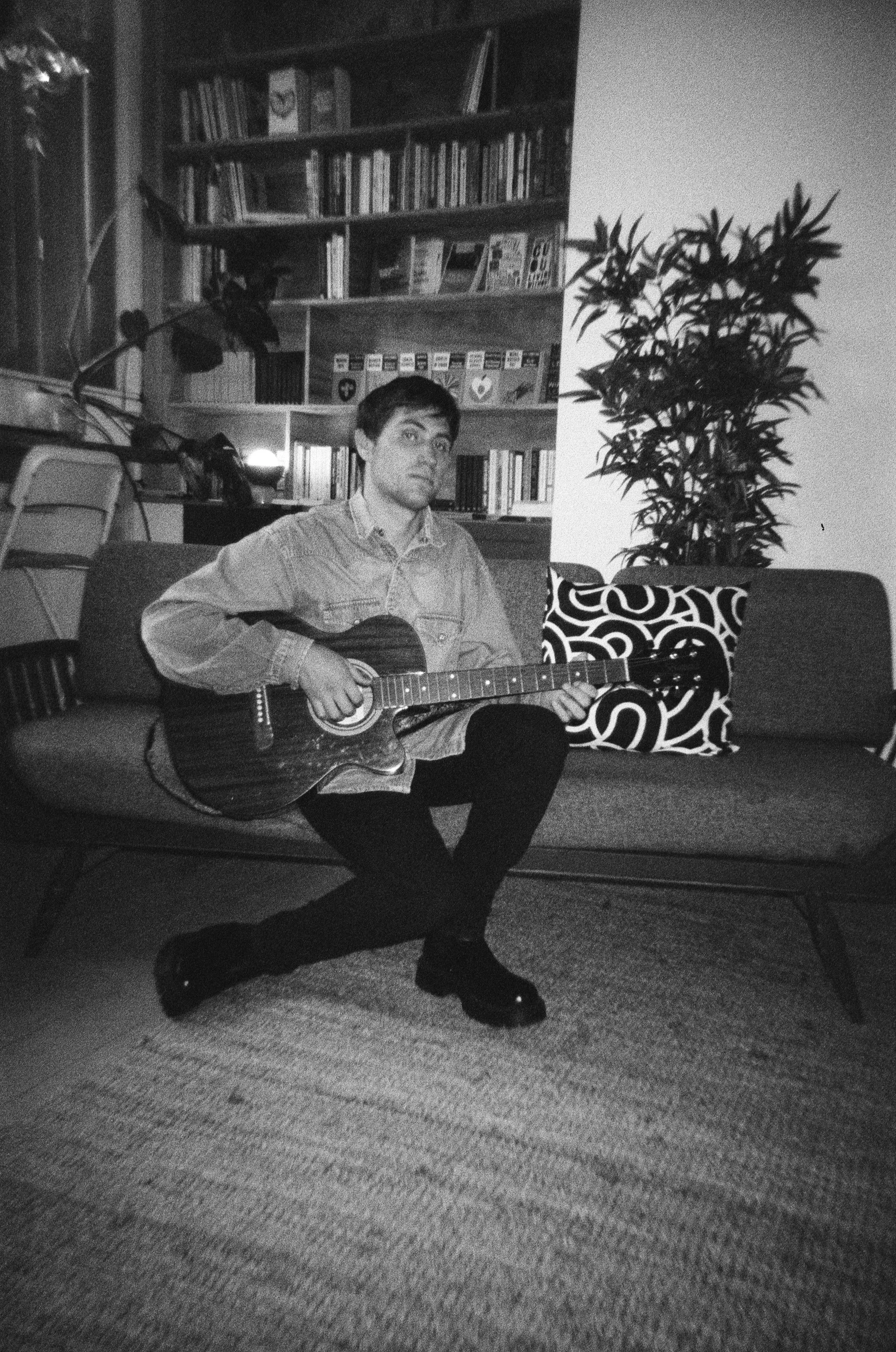 A young man sitting on a sofa with a guitar in a living room, surrounded by bookshelves, a potted plant, and decorative cushions, in black and white photography.