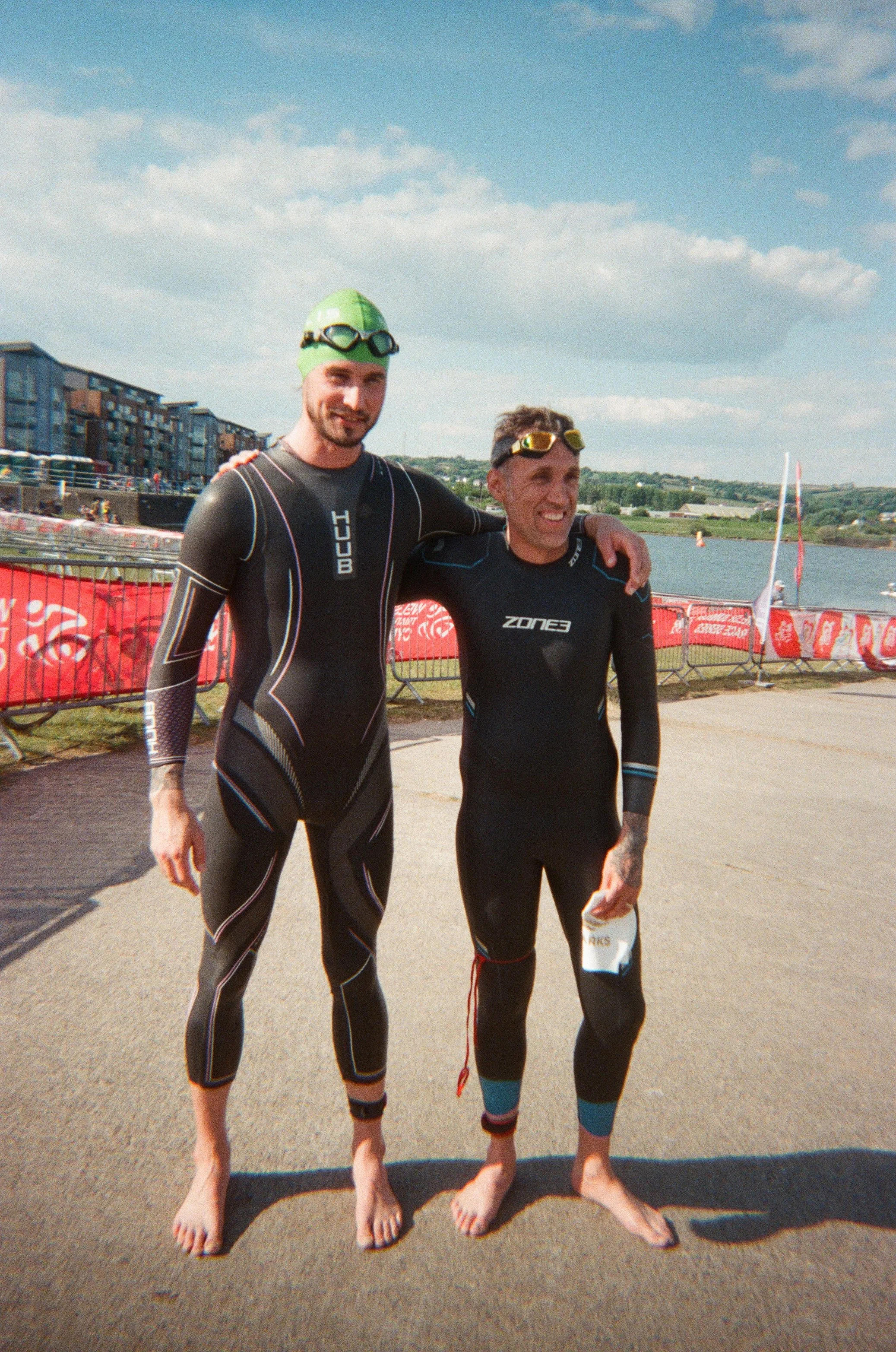 Two men wearing wetsuits and goggles standing on a paved area with a body of water in the background, smiling after a swim or triathlon event.
