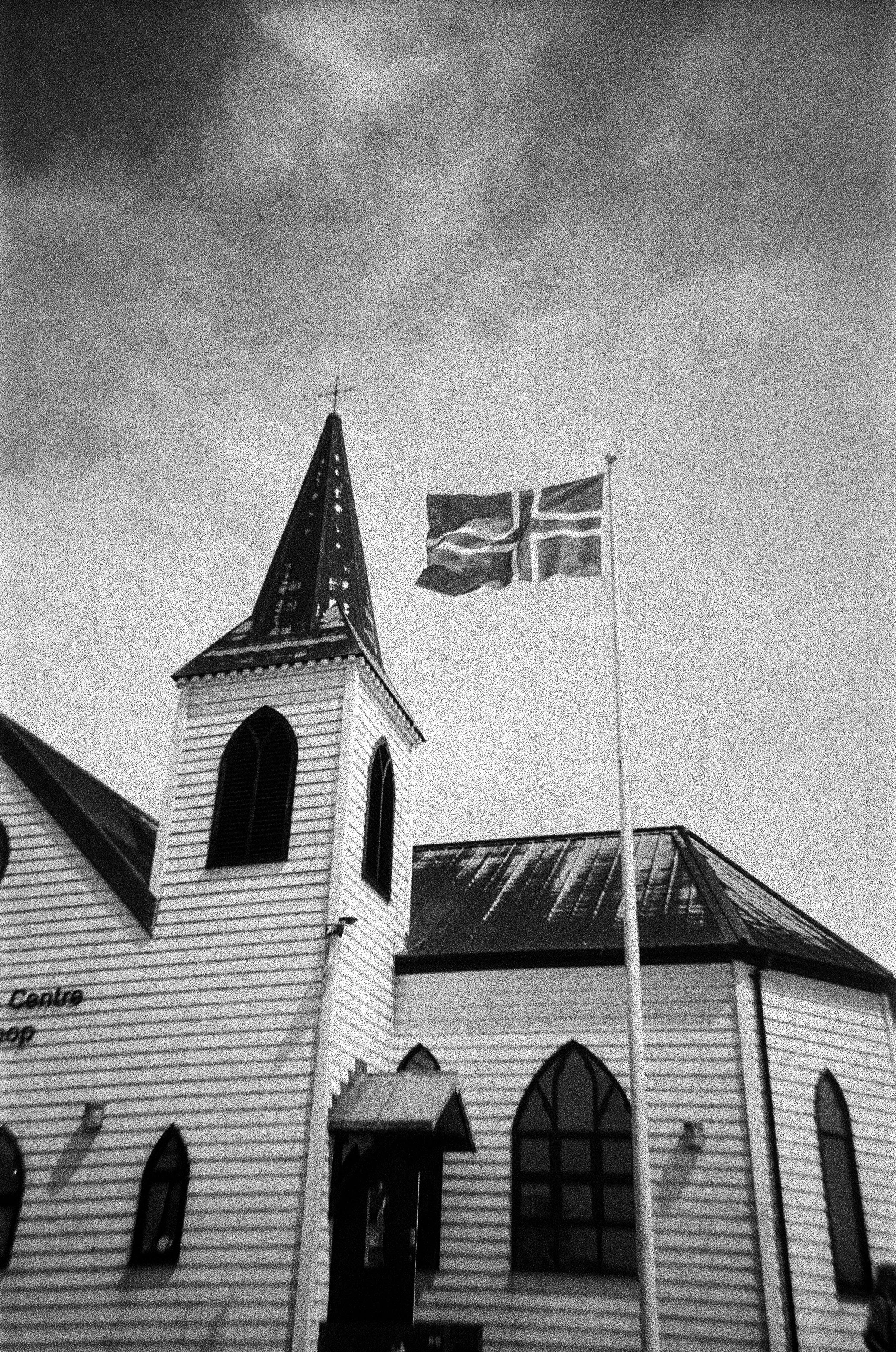 Black and white photo of a church with a steeple, and two flags on a pole in front of it, one of which is the Union Jack.