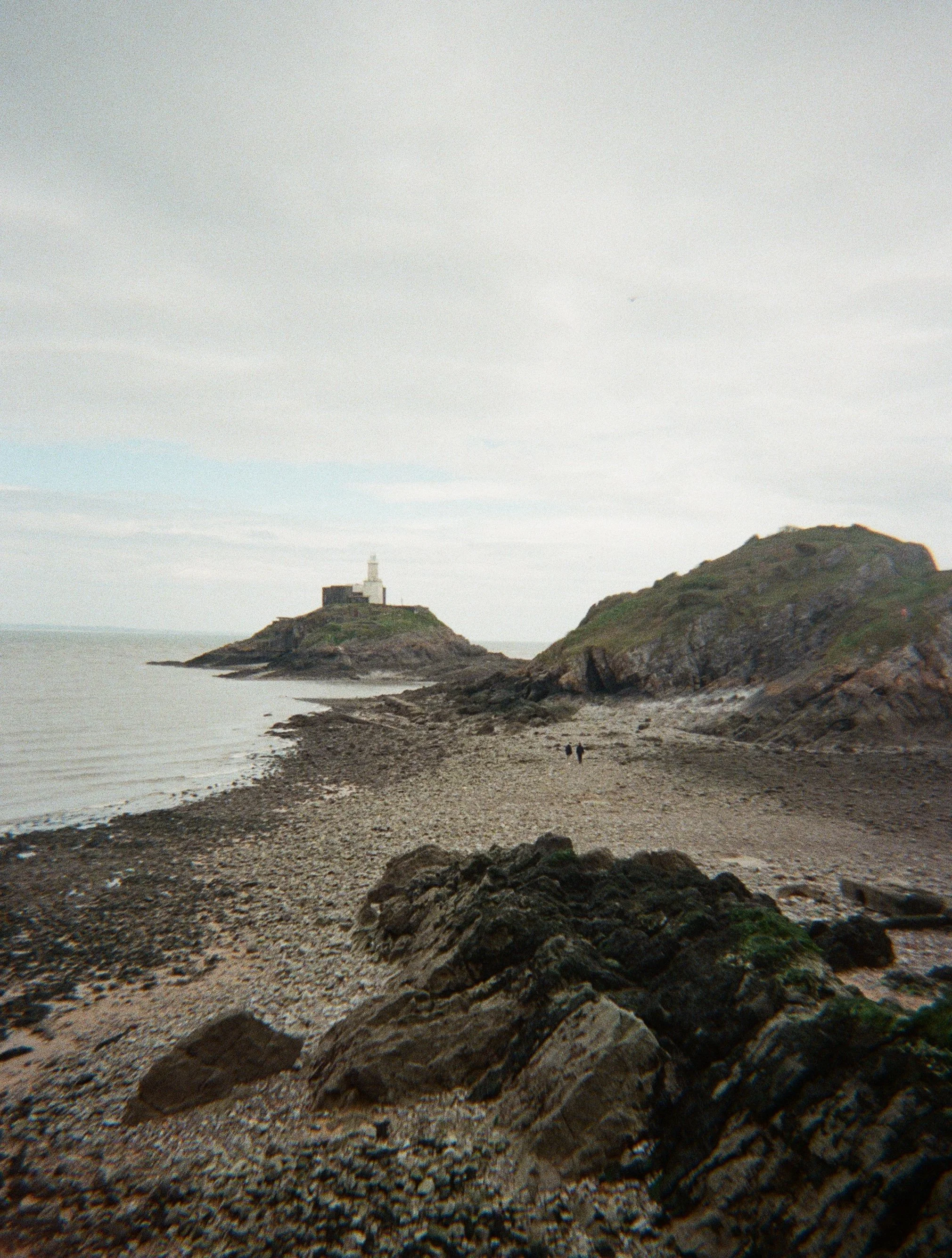 A rocky beach with two people walking towards a hill with a lighthouse on top.