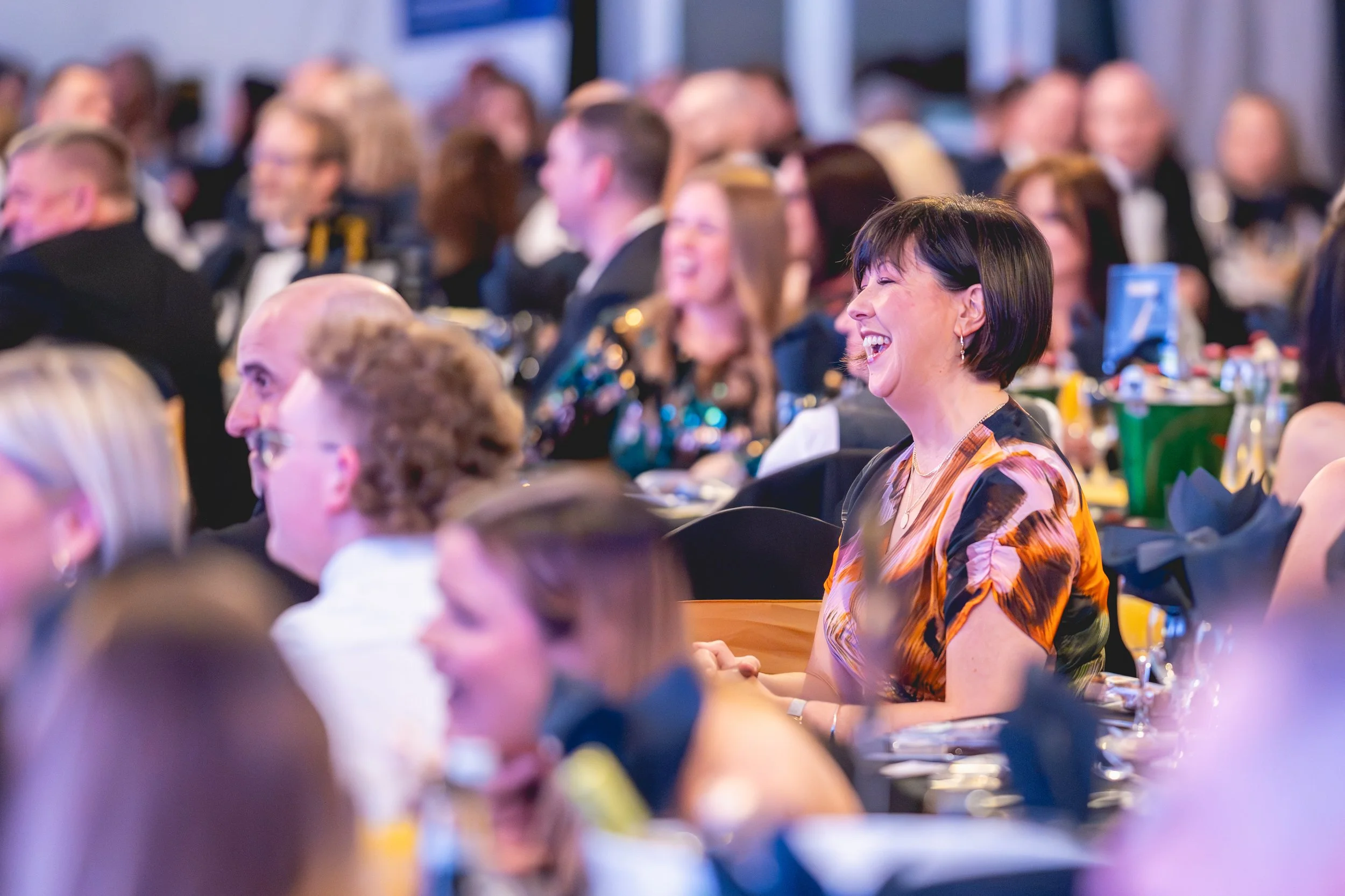 A woman with short black hair wearing a colorful dress, sitting at a formal event, laughing joyfully.