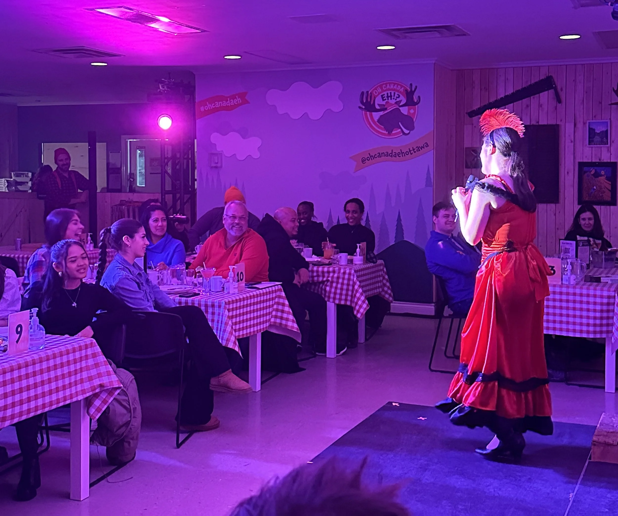A performer dressed in red with a feathered headdress and holding a microphone, performing on a small stage in a restaurant or event space with an audience watching.