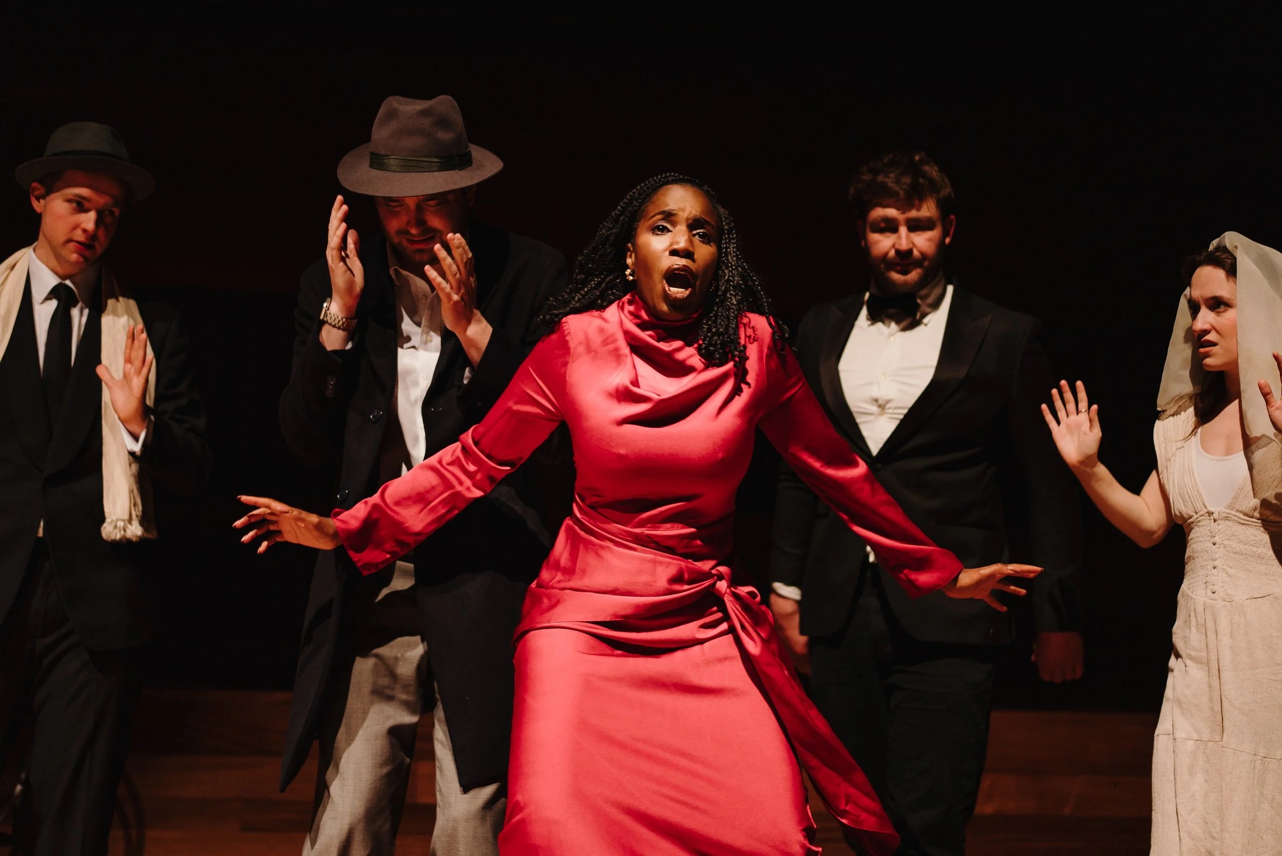 Actors performing on stage, with a woman in a red dress in the center expressing intense emotion, surrounded by performers in vintage costumes.