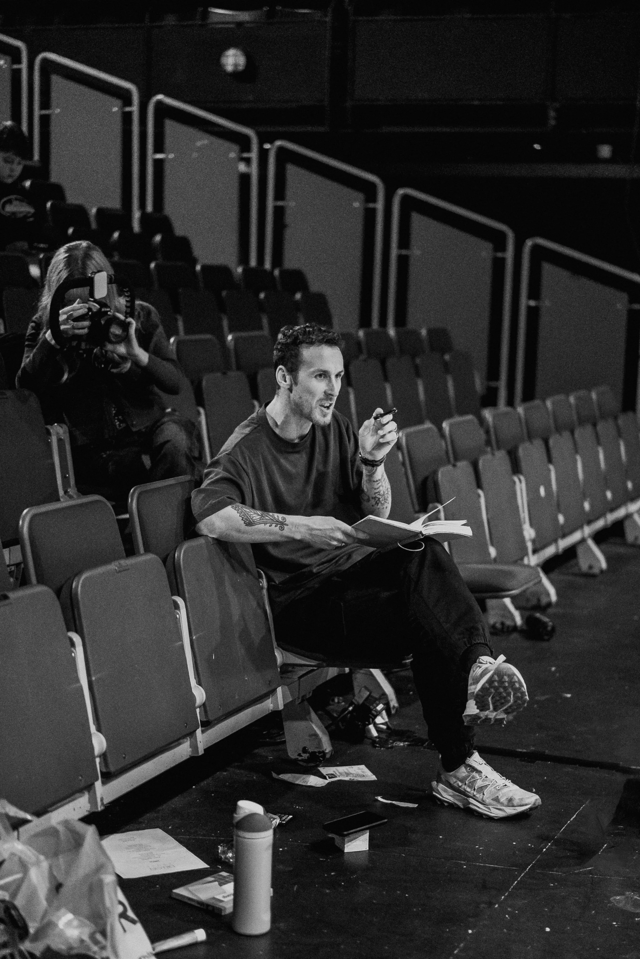 Black-and-white photo of a man with tattoos, sitting in an empty auditorium, speaking into a microphone, with a book in his lap. A woman behind him is taking a photo with a camera.