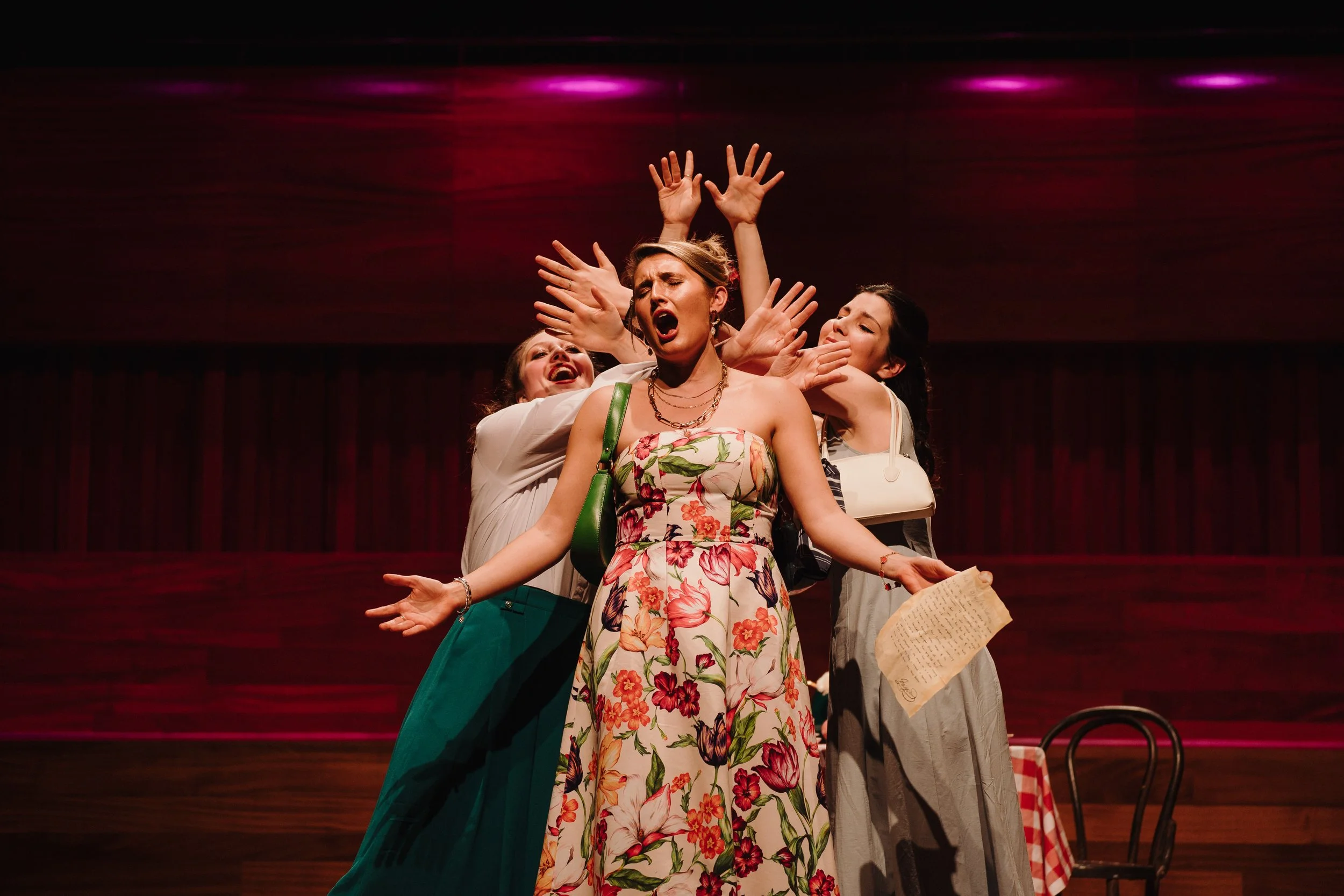 Four women perform on stage with dramatic expressions and gestures, with a dark, wooden background and a single chair to the side.