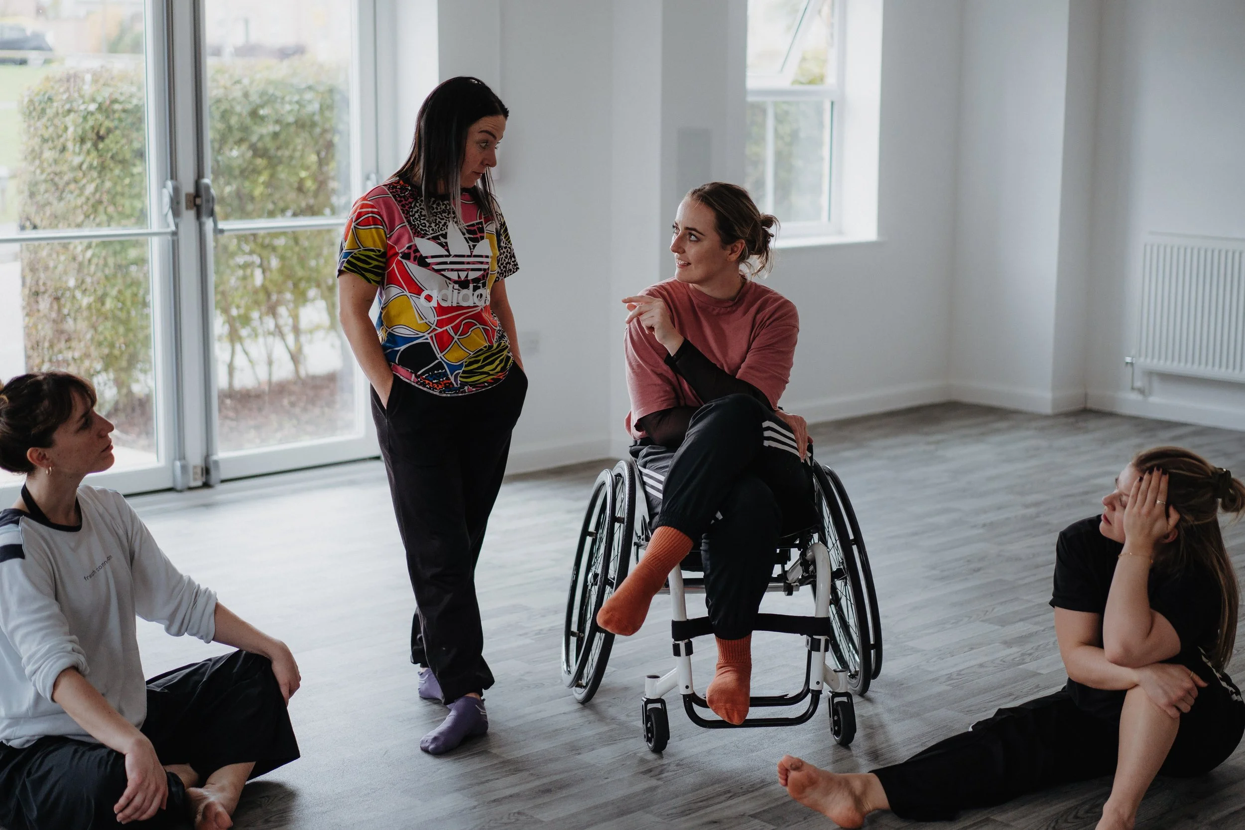 Four women in casual clothing inside a spacious, well-lit room with hardwood floors, engaged in conversation. One woman is seated in a wheelchair, another is sitting on the floor with her head resting in her hand, while the third woman is standing an