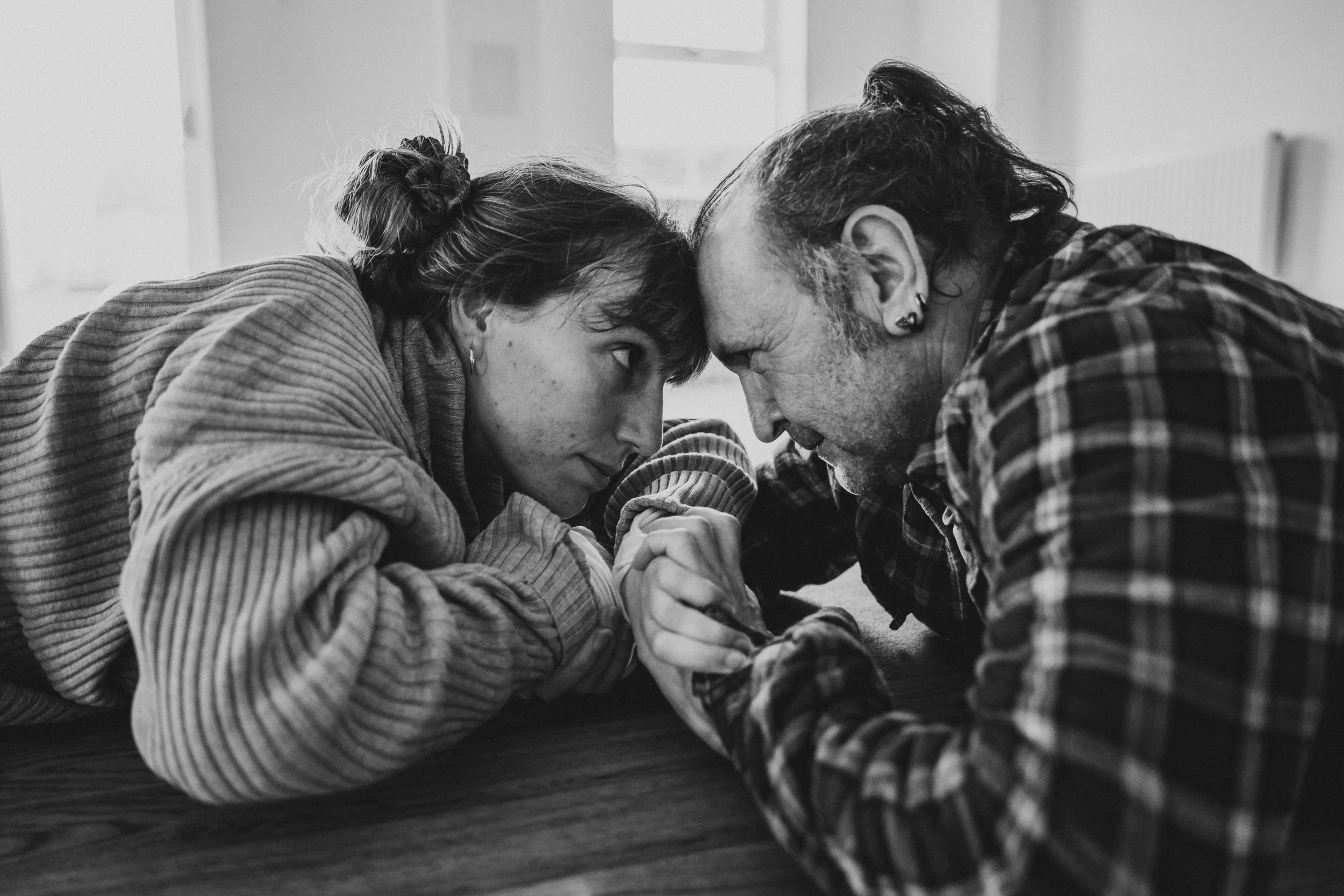 Two people, a woman and a man, are engaged in an arm wrestling match on a table, with their forearms and hands clenched together. They are resting their foreheads against each other, displaying focus and determination. The image is in black and white