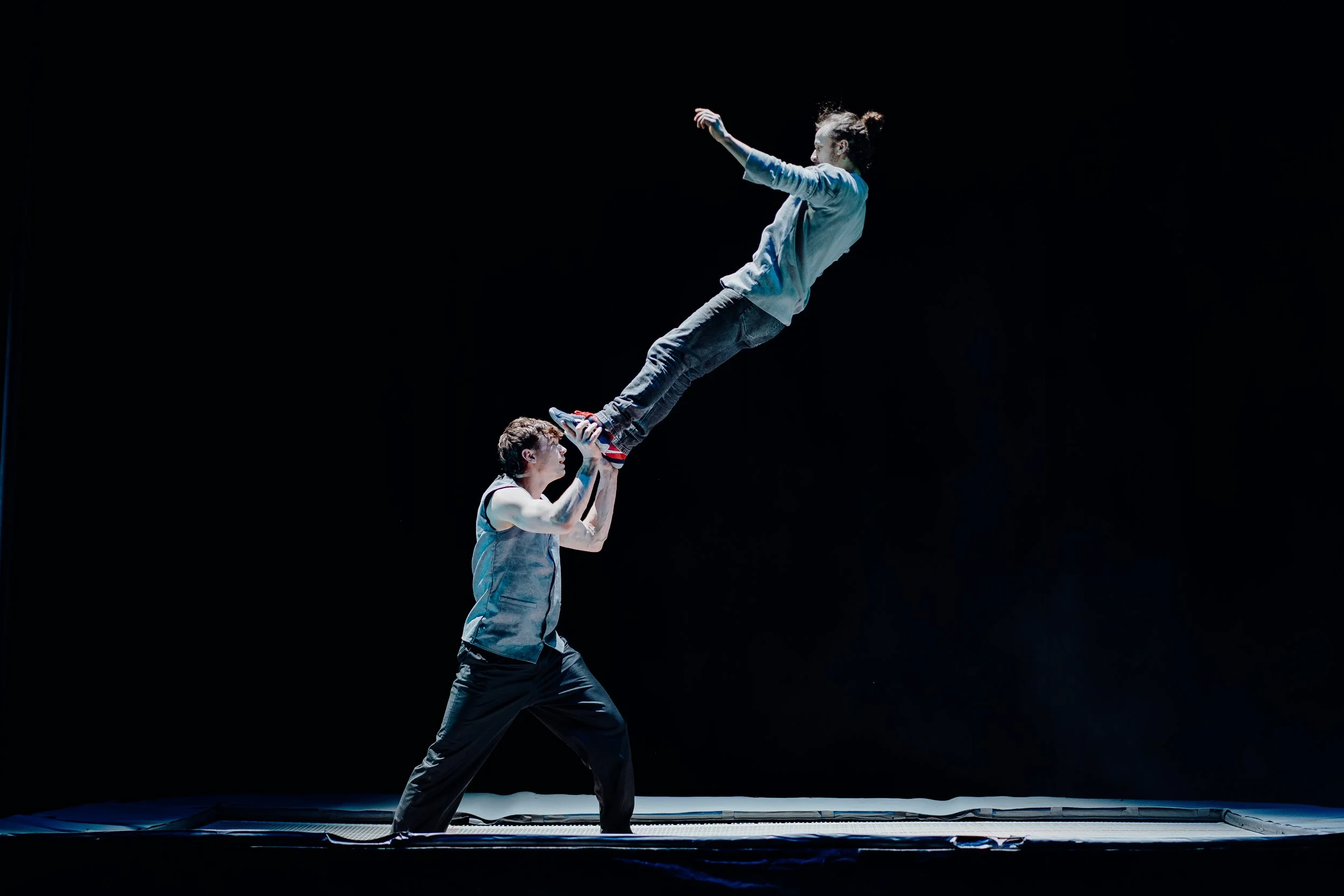Two acrobats performing a balancing act on a trampoline, with one person holding the other upside down above their head, against a dark background.